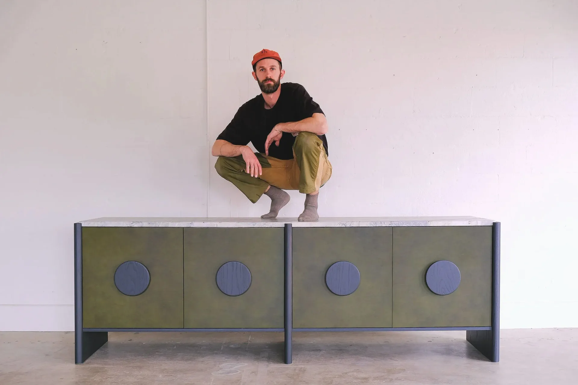 Man in orange beanie crouching on top of a modern two-section credenza with olive green doors and blue circular hardware against a white wall by Scheibe Design.