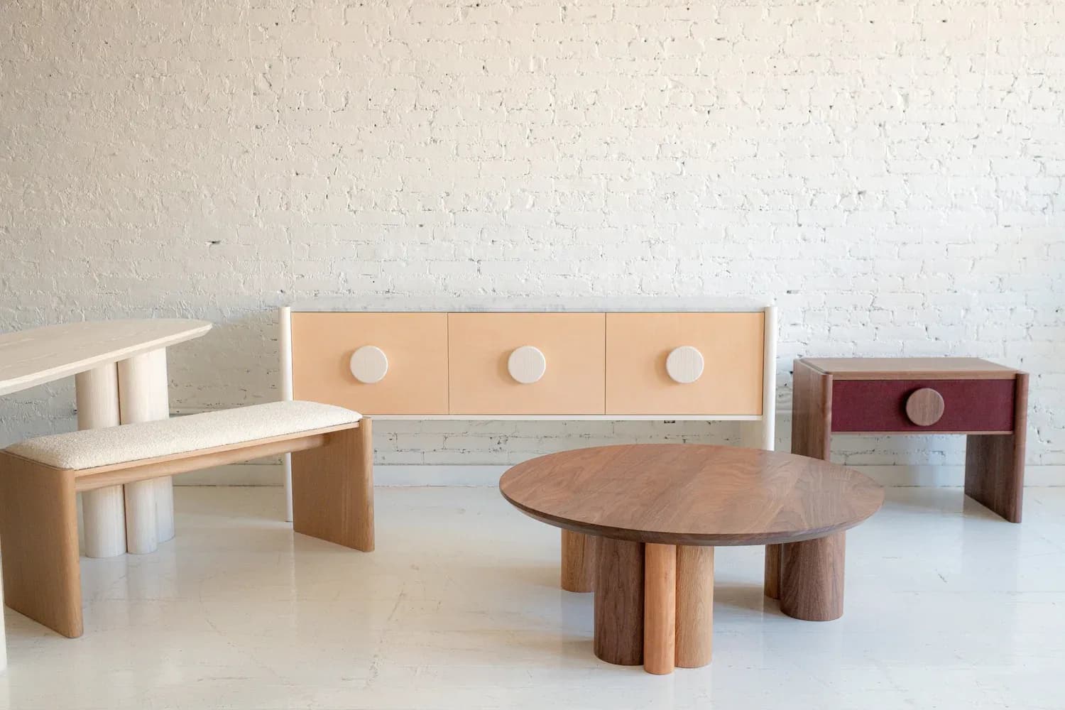 Wooden furniture collection with light natural wood pieces including benches, storage units, and a sculptural oval table with cylindrical legs by Scheibe Design.