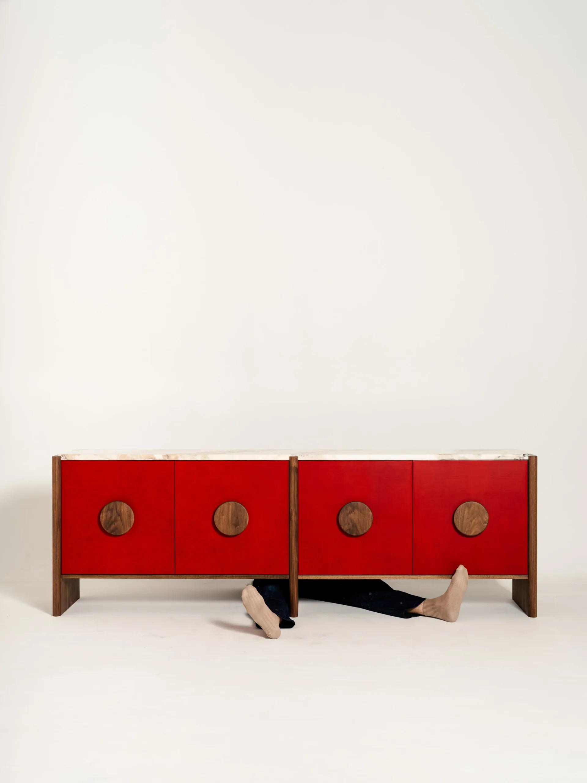 Red lacquered cabinet with four doors and brass circular hardware, featuring wooden frame and legs, with playful sculptural legs emerging underneath, by Scheibe Design.
