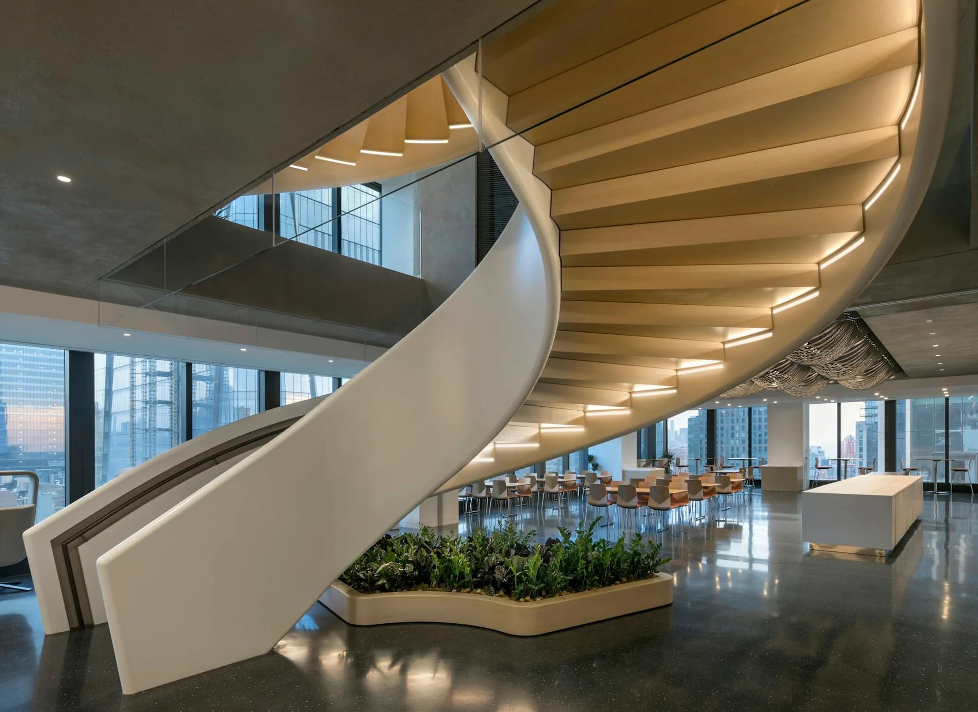Modern spiral staircase with curved white balustrade and integrated warm lighting descends through bright multi-level office space with floor-to-ceiling windows by Schiller Projects.
