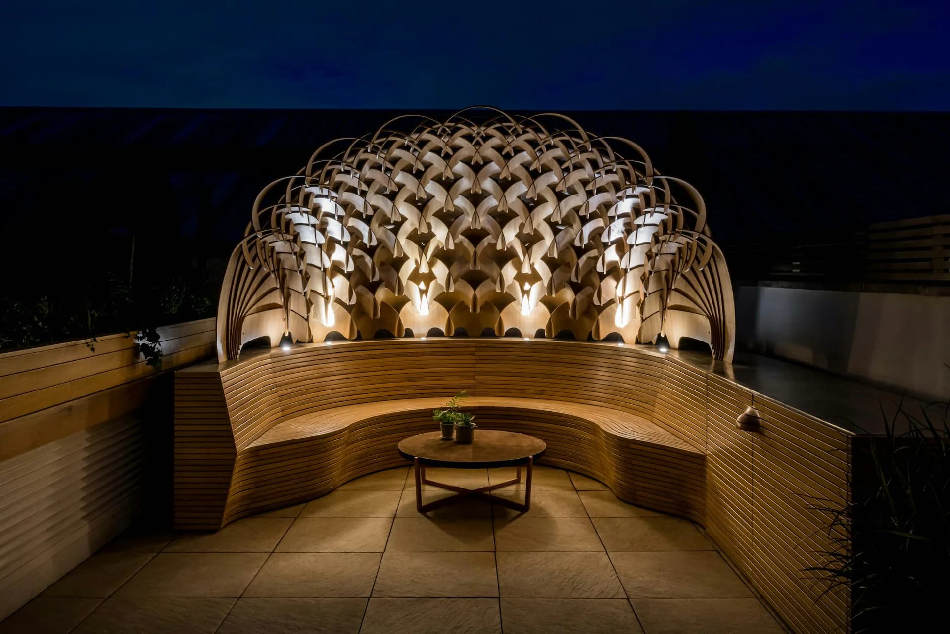 Curved wooden pavilion with illuminated geometric lattice dome structure at night, featuring warm lighting and central seating area by Schiller Projects.