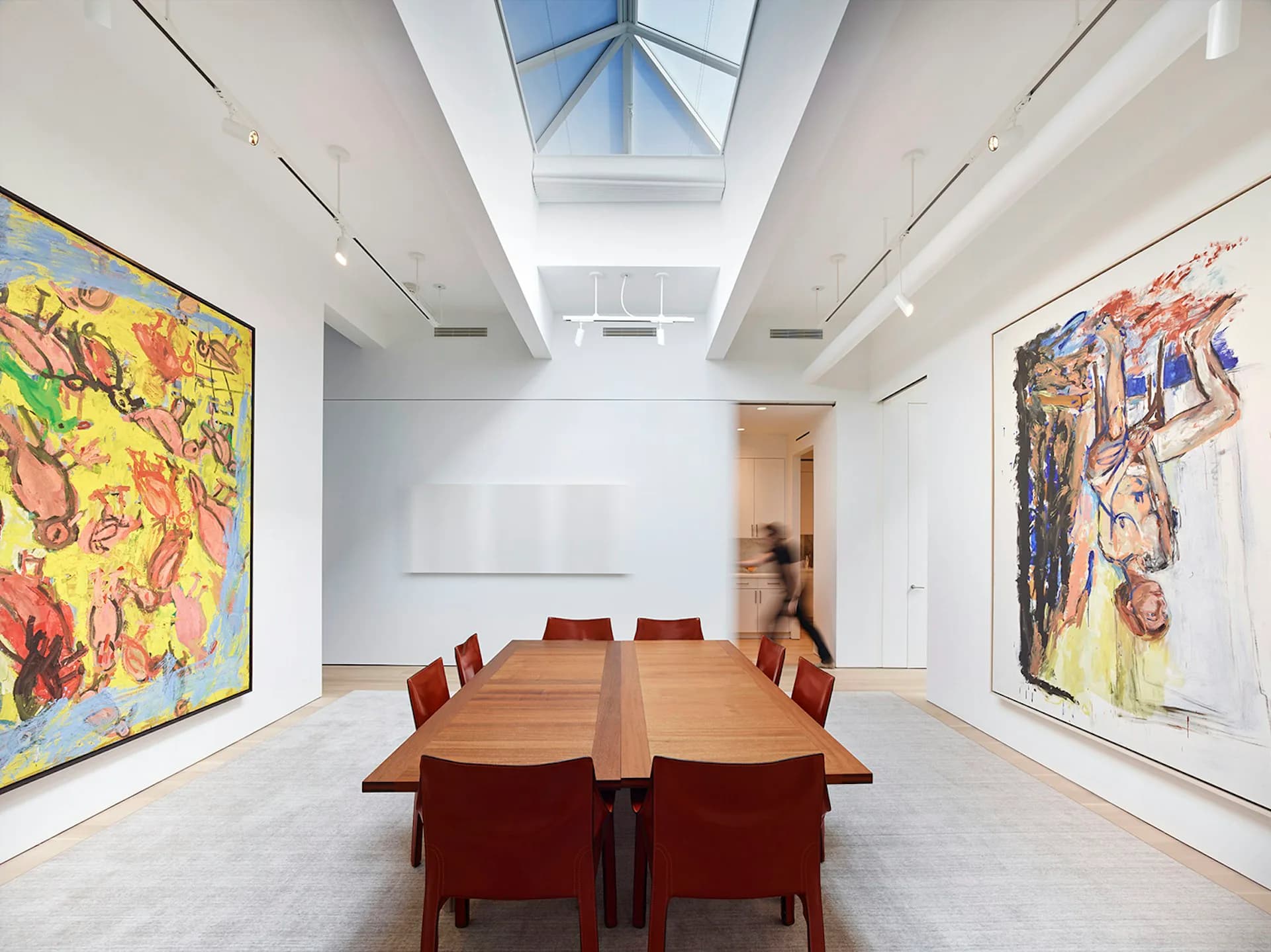 Minimalist gallery dining space with skylight, wooden table, red chairs, and large colorful artworks on white walls by Schiller Projects.