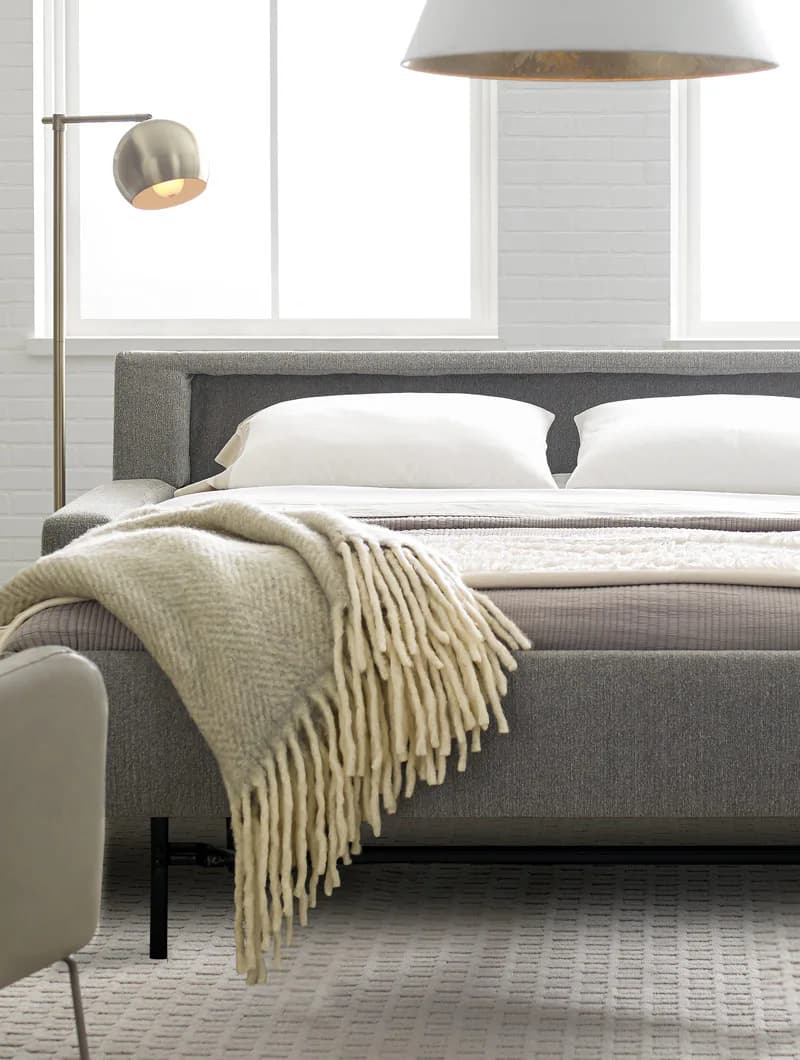 Modern bedroom interior with gray upholstered bed, white pillows, cream throw blanket, pendant and arc floor lamps, floor-to-ceiling windows, and light wood flooring by Scott Jordan Furniture.