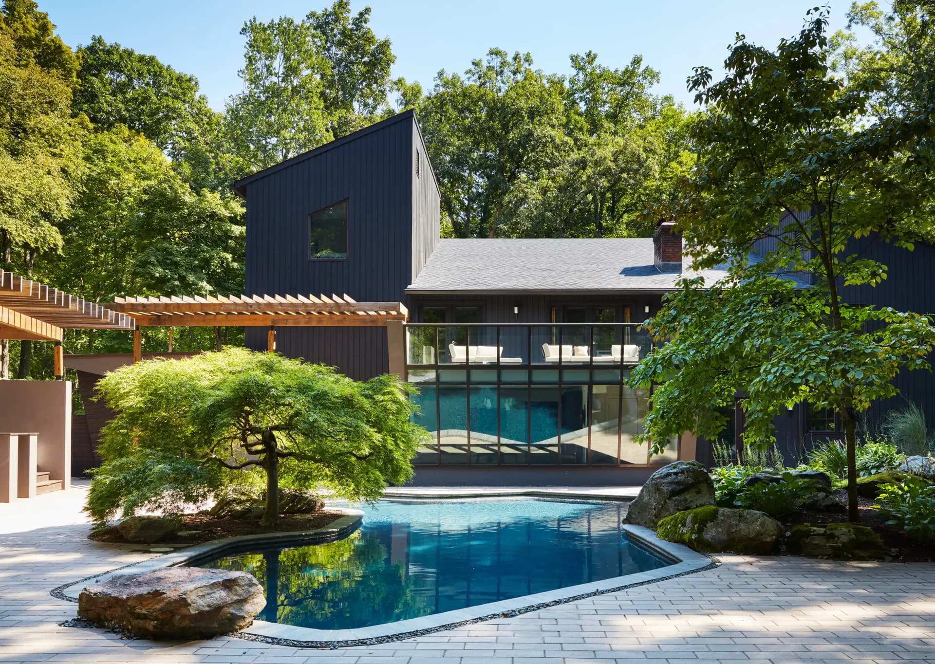 Modern residential pool house with dark metal cladding, glass walls, and integrated swimming pool surrounded by mature trees and manicured landscaping by Scott Springer Architect LLC.
