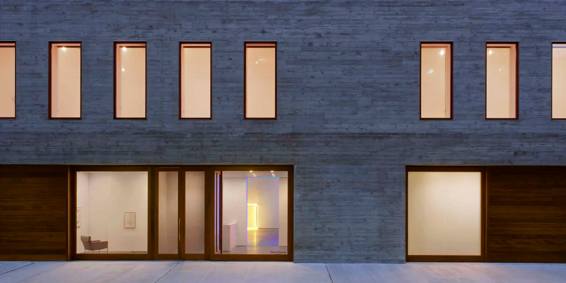 Modern facade with blue-grey brick, horizontal window bands, and dark wood ground-level entries with illuminated interiors by Selldorf Architects.