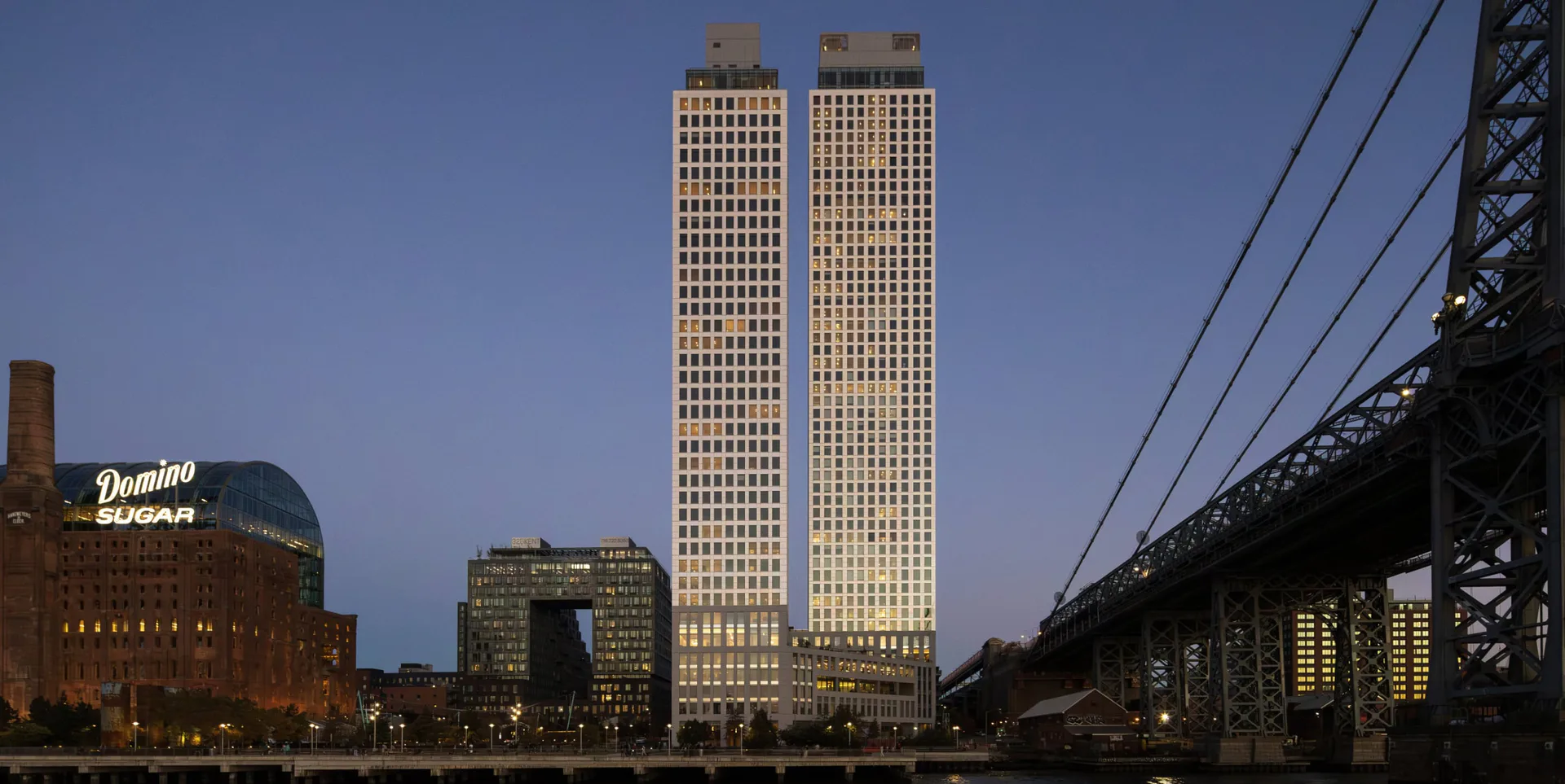 Twilight cityscape featuring two tall rectangular towers with illuminated facades overlooking a waterfront, with bridge infrastructure visible to the right, by Selldorf Architects.