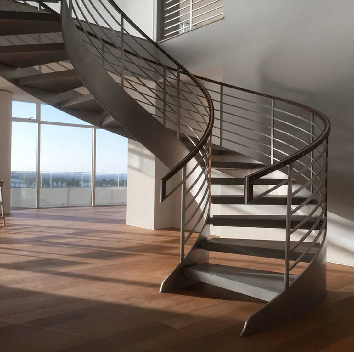 Modern spiral staircase with metal railings in a bright loft space with floor-to-ceiling windows overlooking a cityscape, by Serett Metalworks.