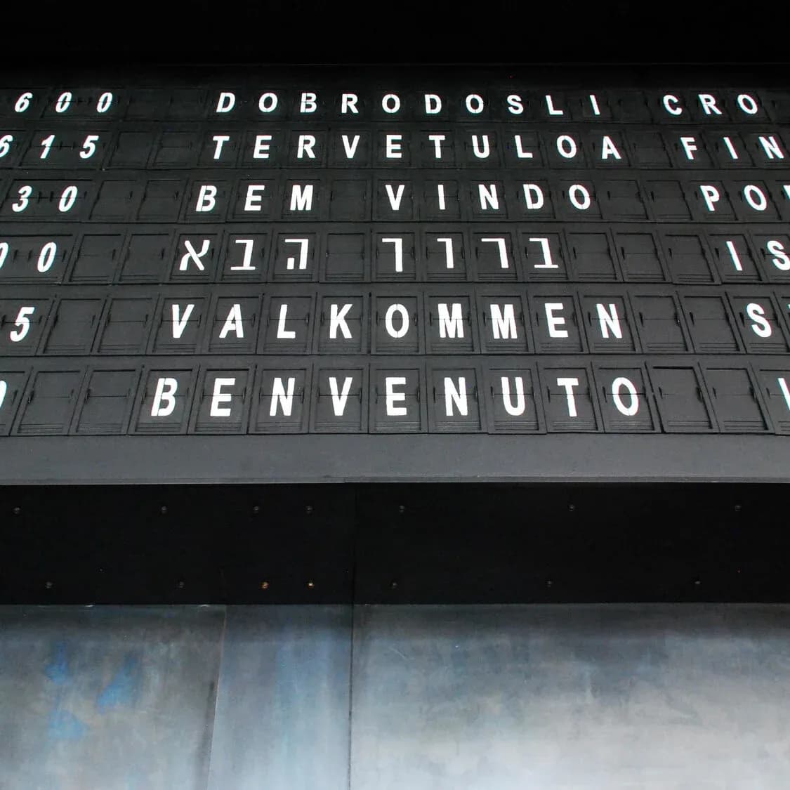 Flip-board airport sign displaying multilingual welcome messages in various languages, featuring bold metalwork lettering by Serett Metalworks.