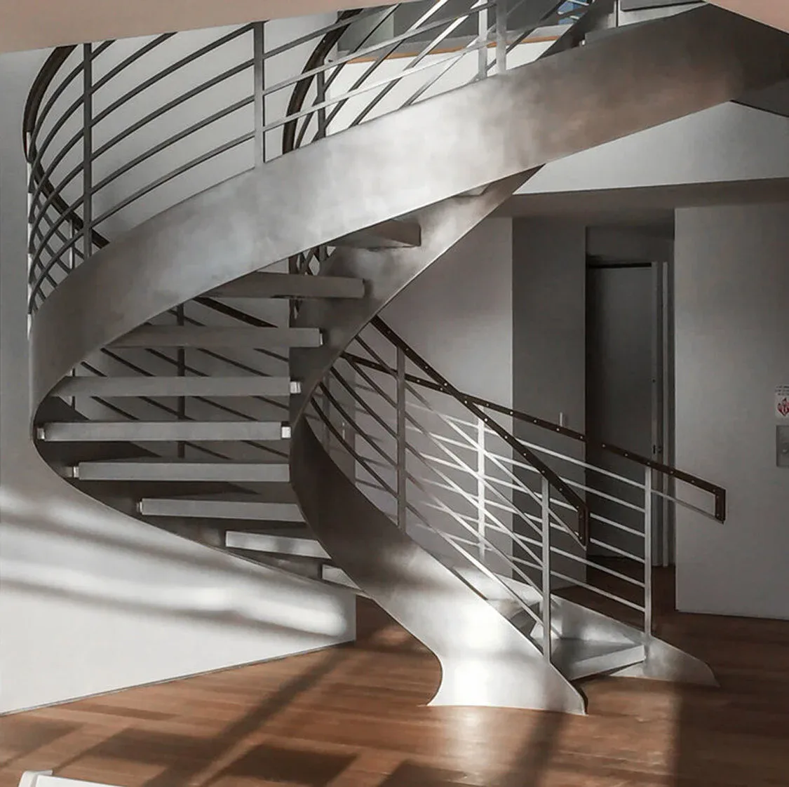 Modern spiral staircase with metal railings and wooden flooring in a bright, minimalist interior by Serett Metalworks.