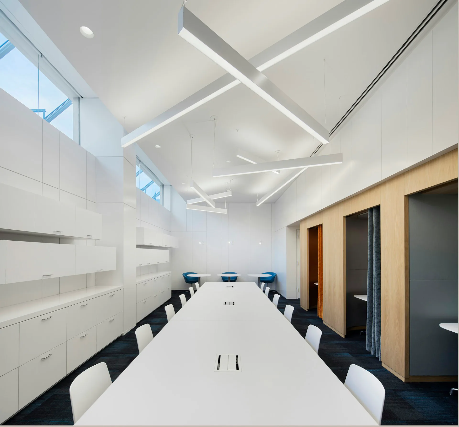 Modern conference room with white ceiling, skylights, long white table, black chairs, wooden accent doors, and built-in cabinetry by Shakespeare Gordon Studio.