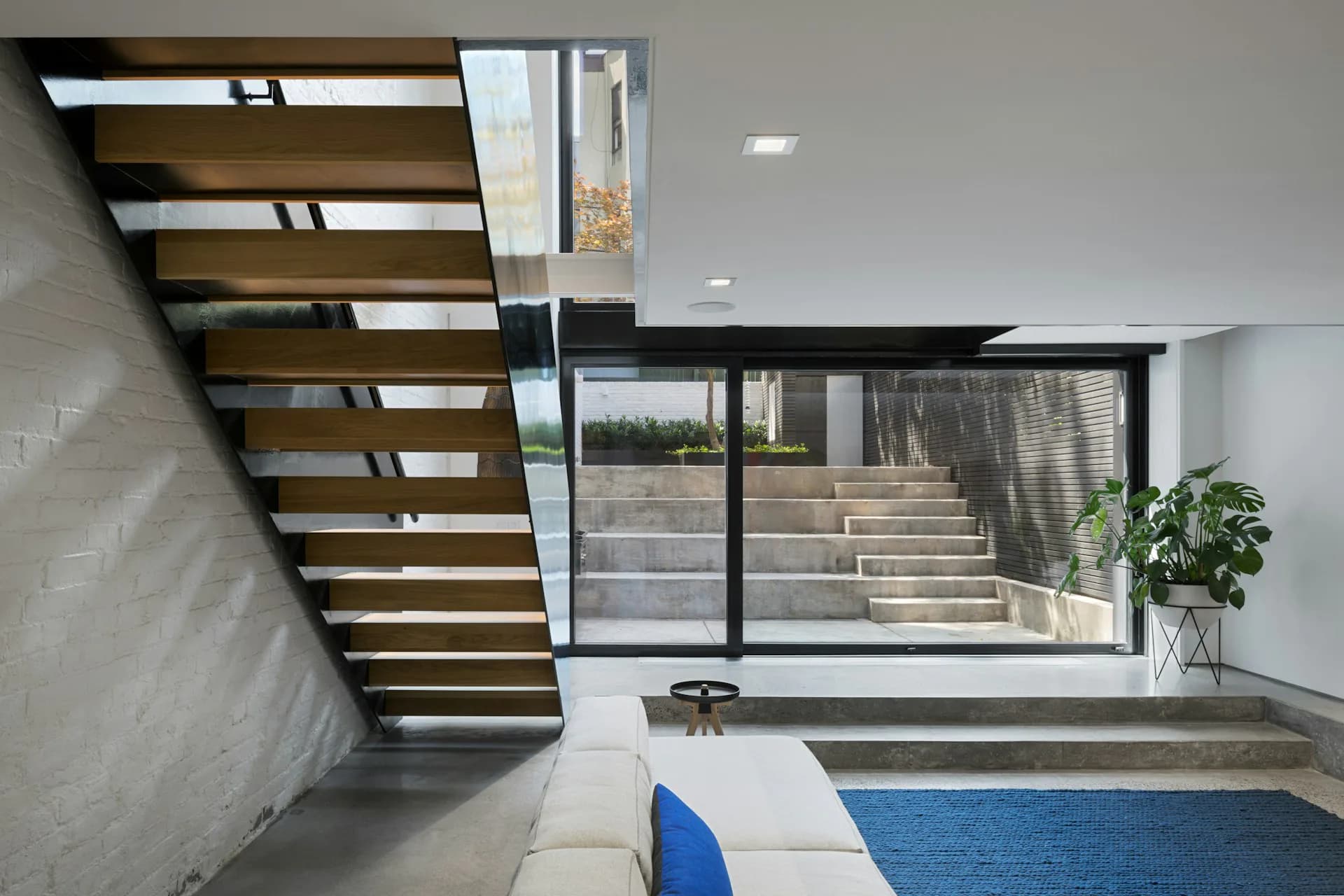 Modern basement interior with floating wooden staircase, polished concrete floors, blue accent rug, and large window overlooking outdoor stairs by Shakespeare Gordon Studio.