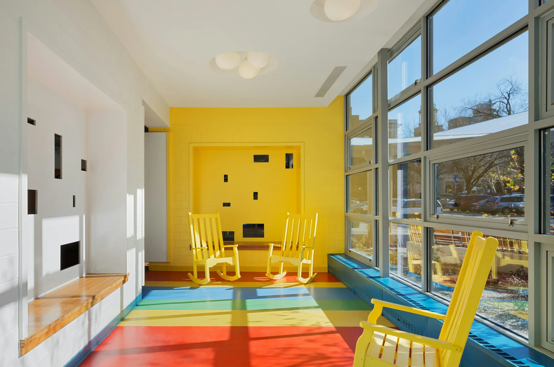 Bright residential interior with yellow accent wall, colorful striped floor, and large windows overlooking landscape by Shakespeare Gordon Studio.