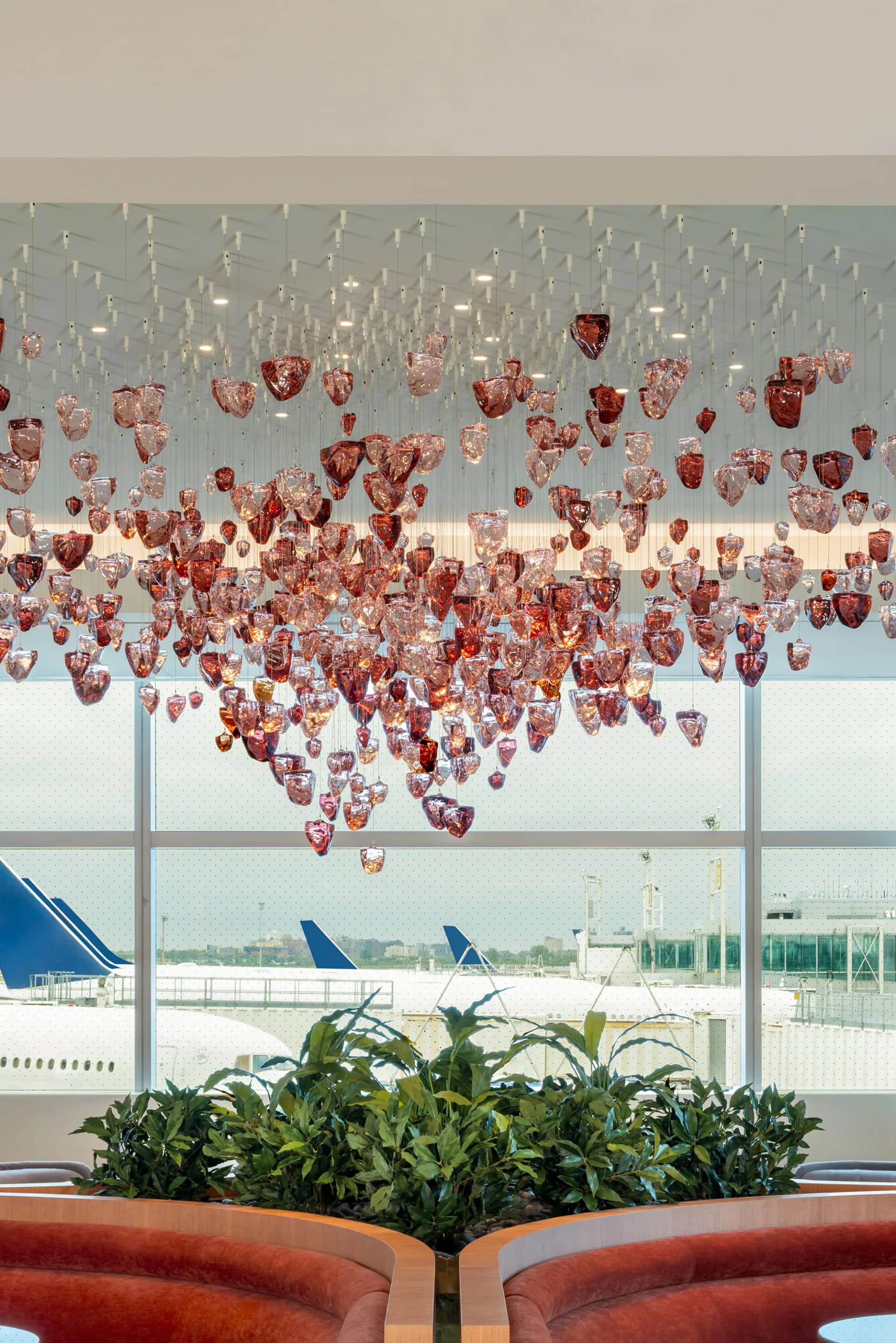 Airport terminal interior with suspended red heart-shaped decorative installation above green plants, overlooking tarmac and aircraft, by Shakúff.