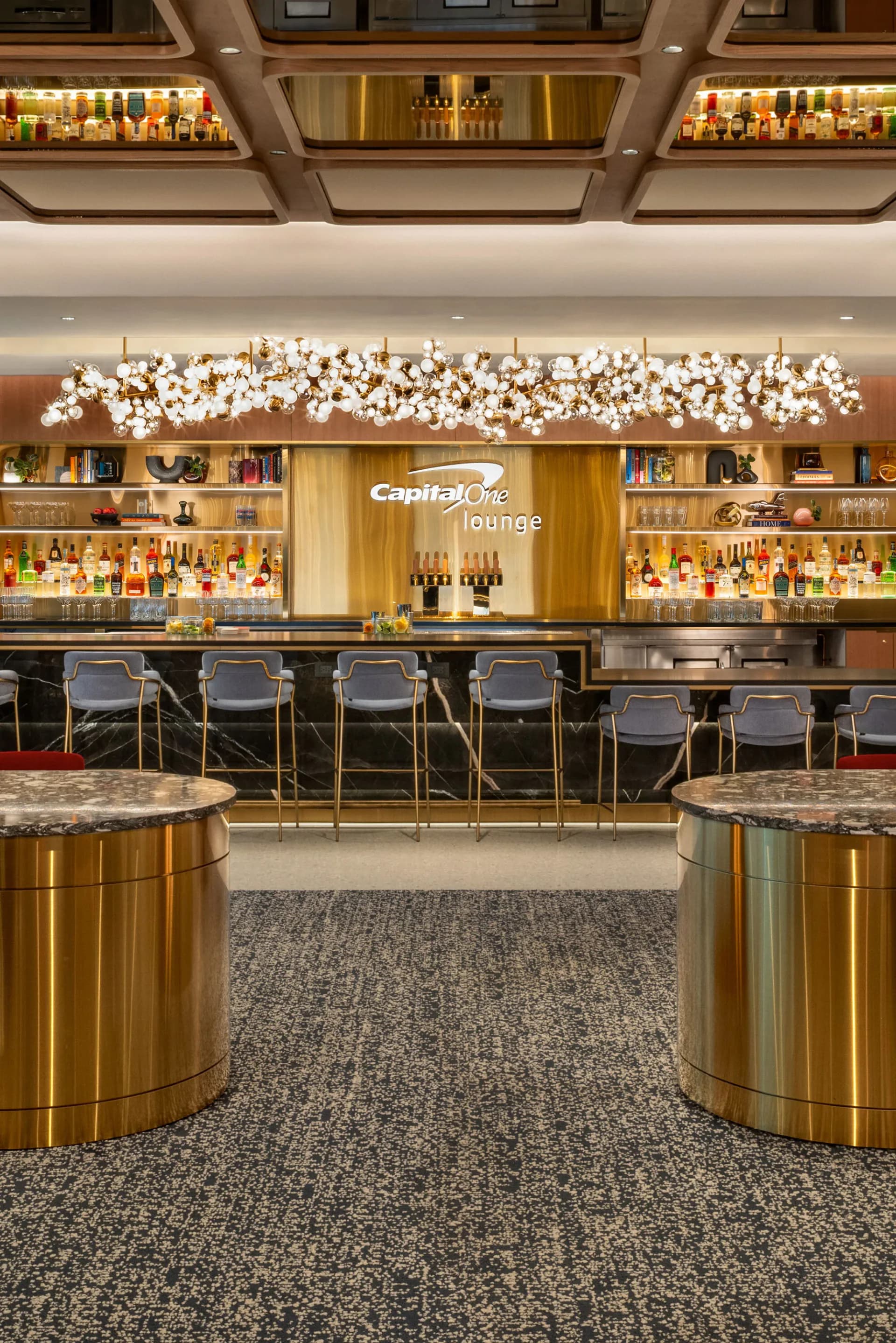 Capital One Lounge bar interior featuring brass-accented details, illuminated cloud-like pendant installation, extensive liquor shelving, and gray terrazzo flooring by Shakuff.