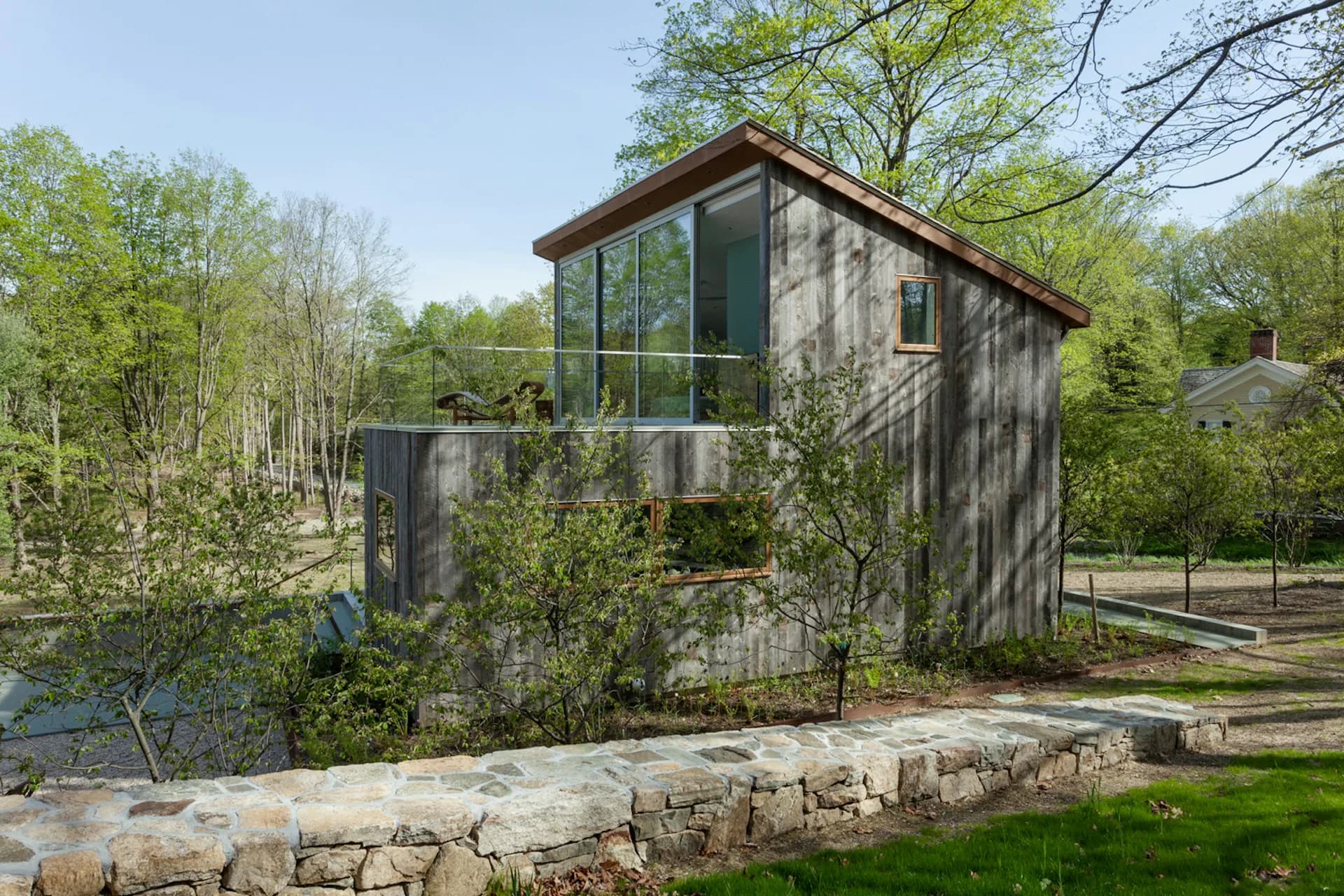 Modern dark-clad residential structure with extensive glazing overlooking a wooded landscape and stone retaining wall by Sharon Davis Design.