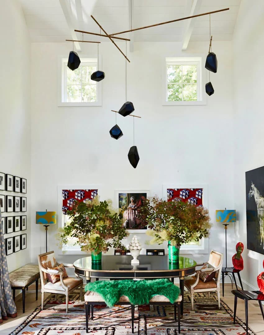 Modern living room with black oval dining table, woven chairs, green plant arrangements, brass mobile sculpture, and contemporary artwork on white walls by Sheila Bridges Design.