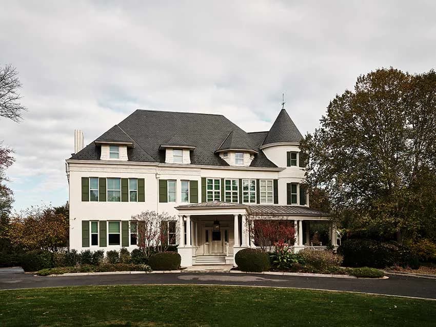 A grand Victorian-style white exterior with mansard roof, multiple dormers, and green shutters, surrounded by manicured landscaping by Sheila Bridges Design.