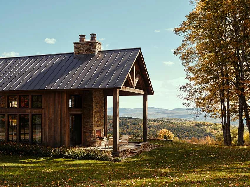 Modern farmhouse with dark metal roof and stone exterior overlooks a scenic lake and forested valley, combining rustic and contemporary architectural elements by Sheila Bridges Design.