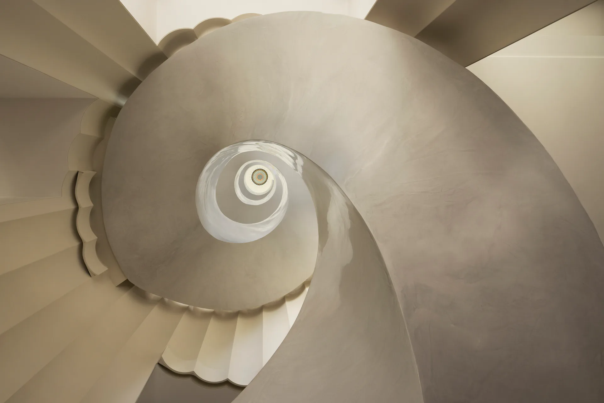 Spiral staircase with cream-colored curved walls and steps photographed from above, creating a geometric pattern of descending circular forms by SheltonMindel℠.