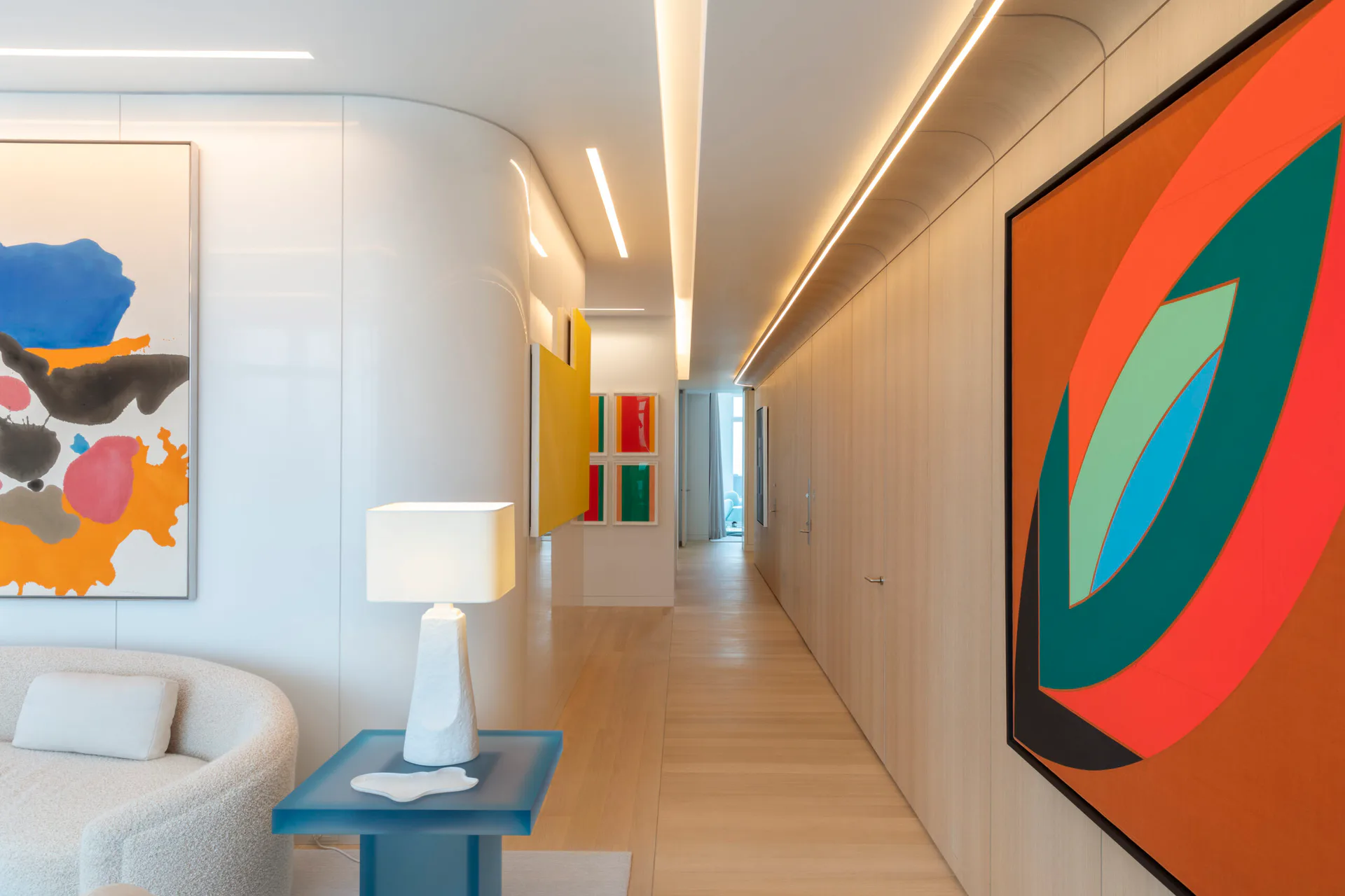 Modern corridor with curved white walls, warm linear ceiling lights, colorful abstract artworks, and polished wood floors, designed by SheltonMindel℠.