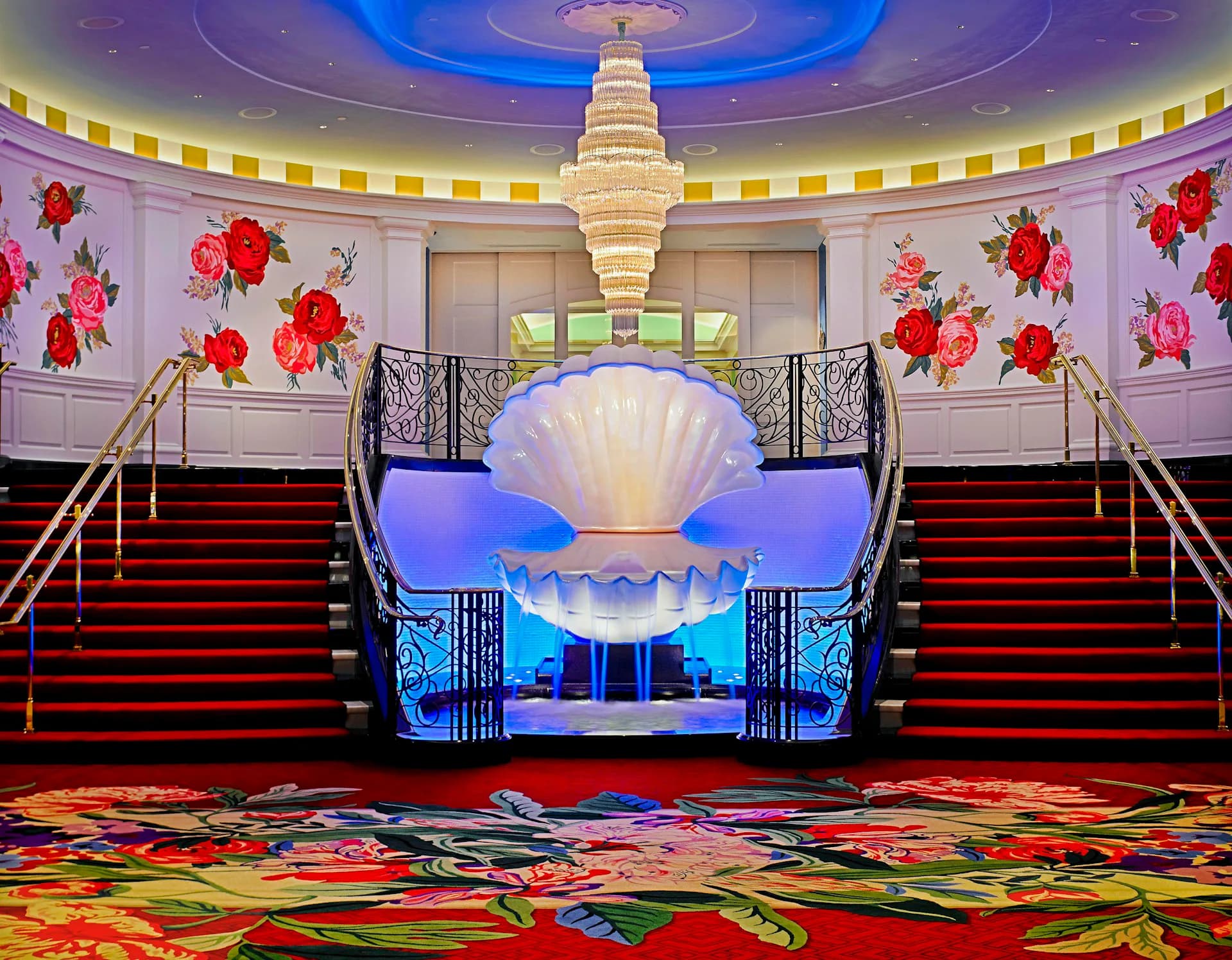Grand hotel lobby with symmetrical red-carpeted staircase, illuminated shell-shaped backdrop, hanging crystal chandelier, and red floral wall installations by Shope Reno Wharton Associates.