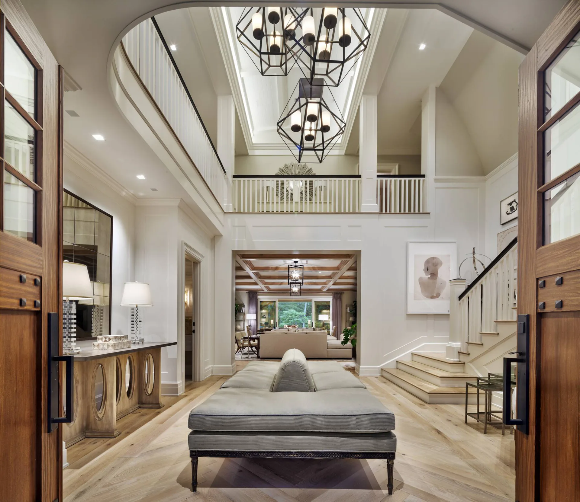 A two-story foyer with soaring ceilings, geometric pendant lights, wood-paneled doors, floating staircase, and polished stone flooring in warm neutral tones by Shope Reno Wharton Associates.