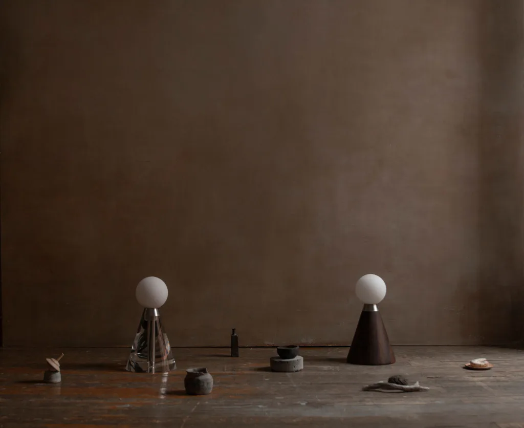 Minimalist tabletop installation featuring two cone-shaped black vessels with white spheres on top, arranged with small sculptural objects on a weathered wooden surface against a neutral brown wall by Siete Studio.