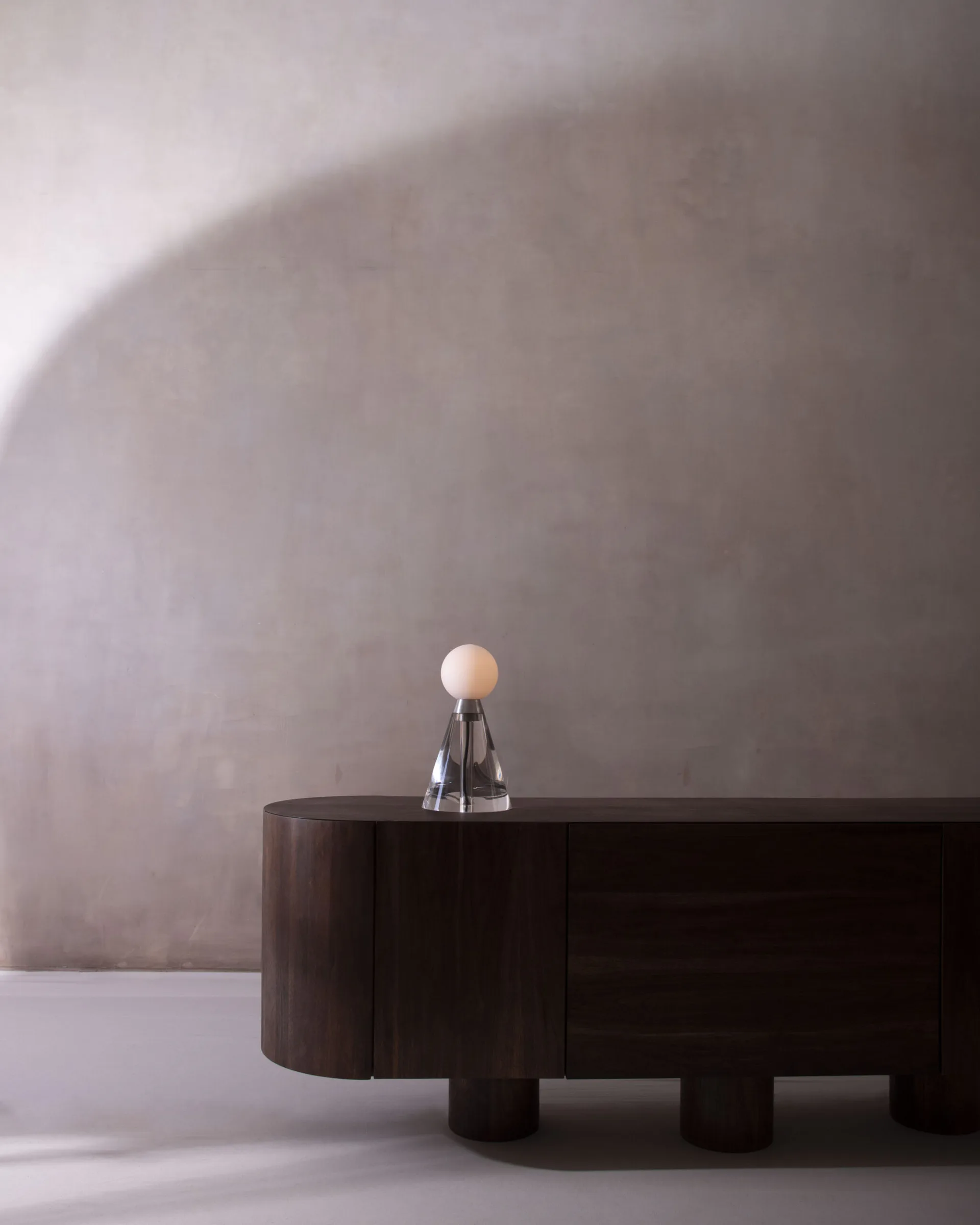 Minimalist interior with dark curved table and sculptural white sphere on pedestal against textured wall by Siete Studio.