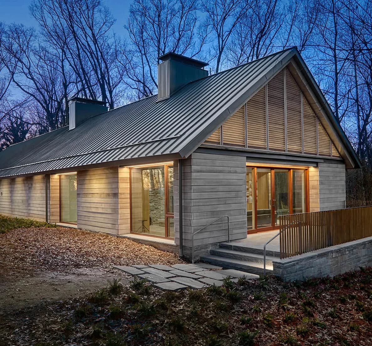 Modern residential home with charcoal siding and metal roof features large glazed gable end and wooden deck, surrounded by bare trees by Sighte Studio.