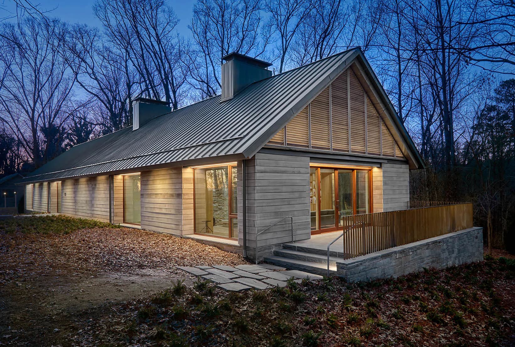 Modern residential house with board-and-batten siding, metal roof, and large glazed openings illuminated at dusk, surrounded by bare trees, by Sighte Studio.