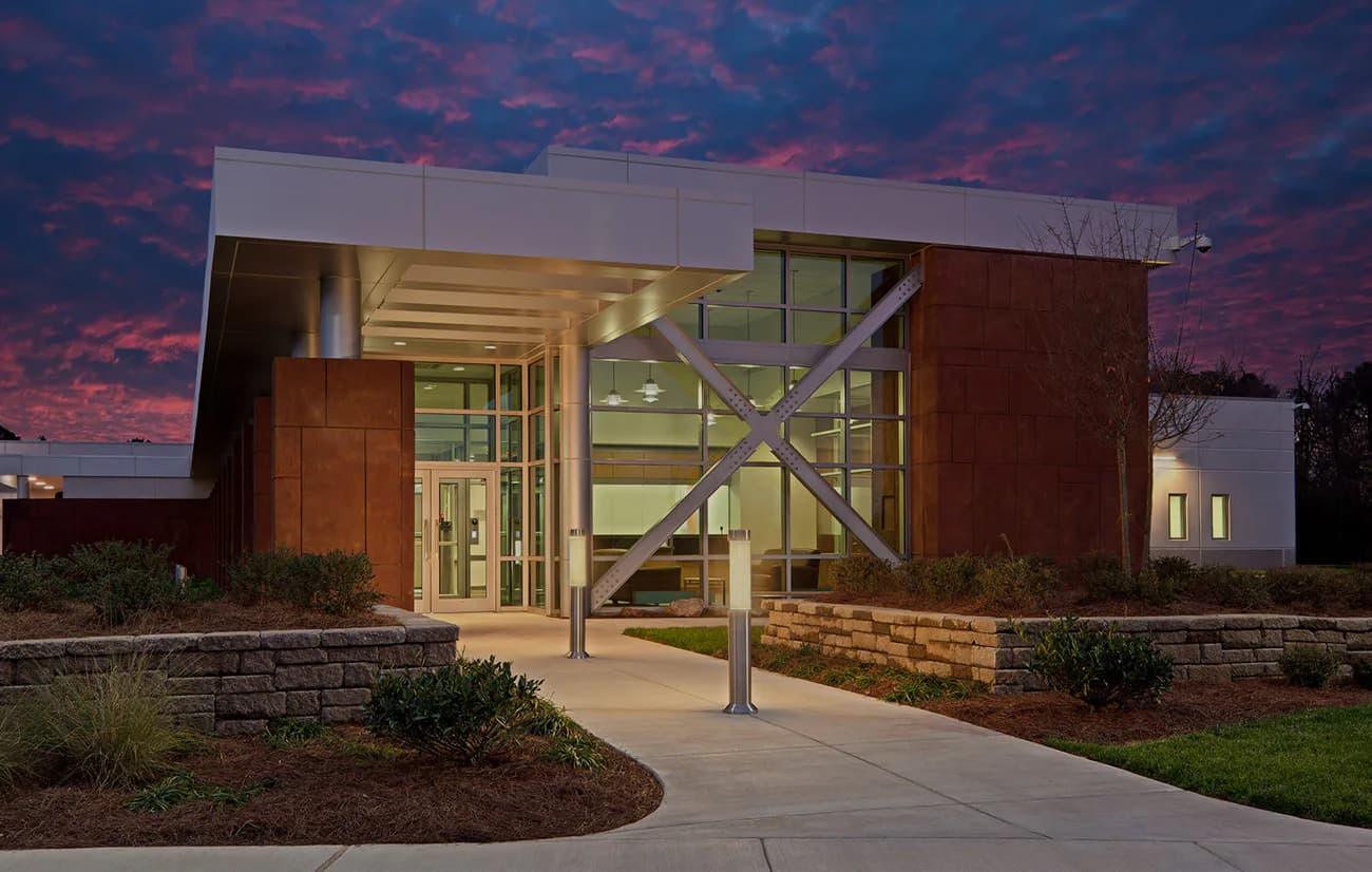Modern institutional building with warm wood and metal cladding, illuminated entrance, and stone base photographed at dusk by Sigma7 design group.