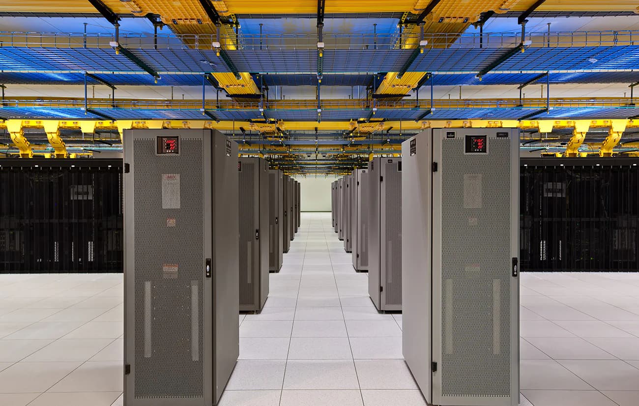 Data center corridor with gray server cabinets and yellow overhead cable management system on white flooring by Sigma7 design group.