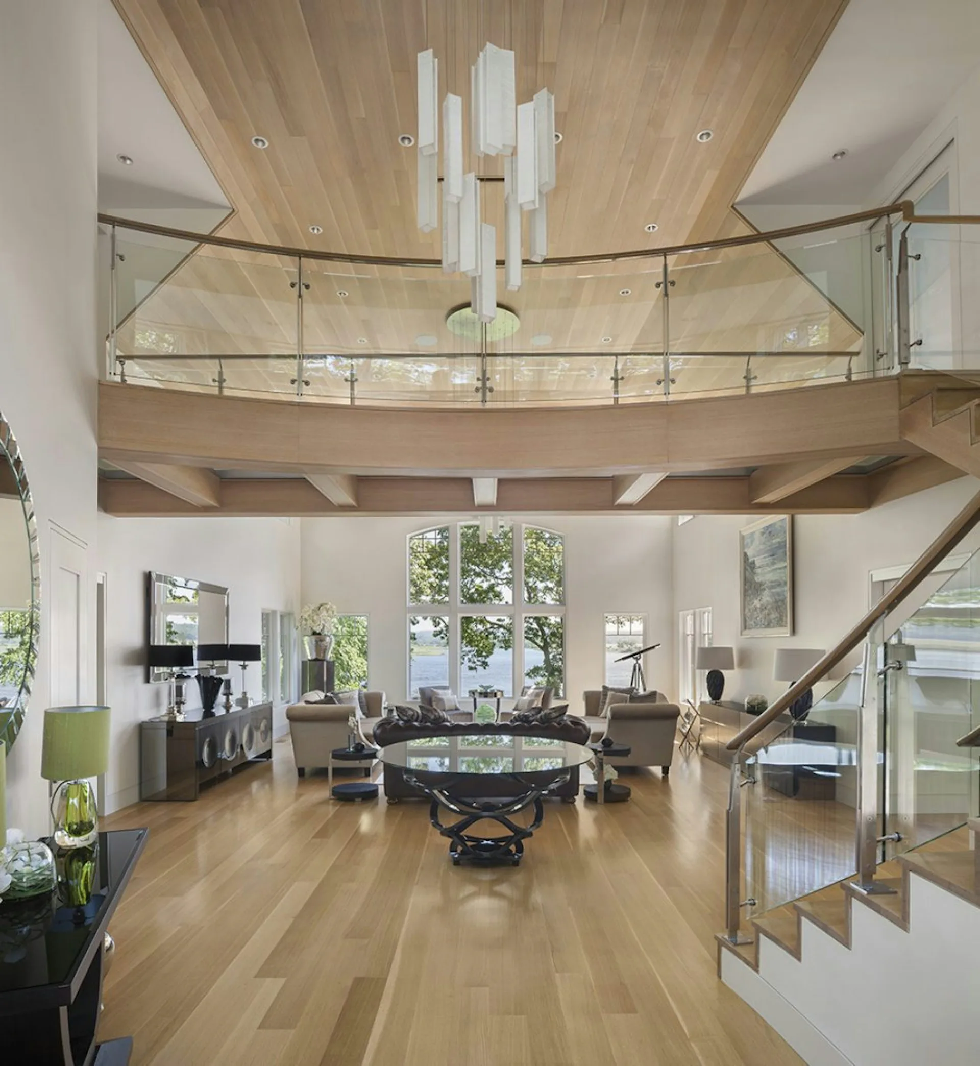 Modern two-story open-plan living space with light wood ceiling, glass railings, white pendant lights, and warm wood flooring by Siris Coombs Architecture.