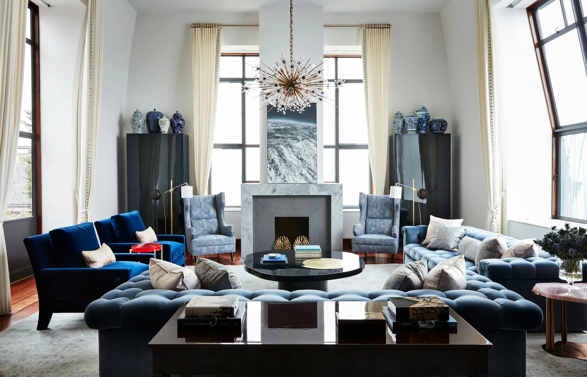 Symmetrical living room with soaring ceilings, black velvet seating, marble fireplace, blue accents, and brass chandelier by Siris Coombs Architecture.