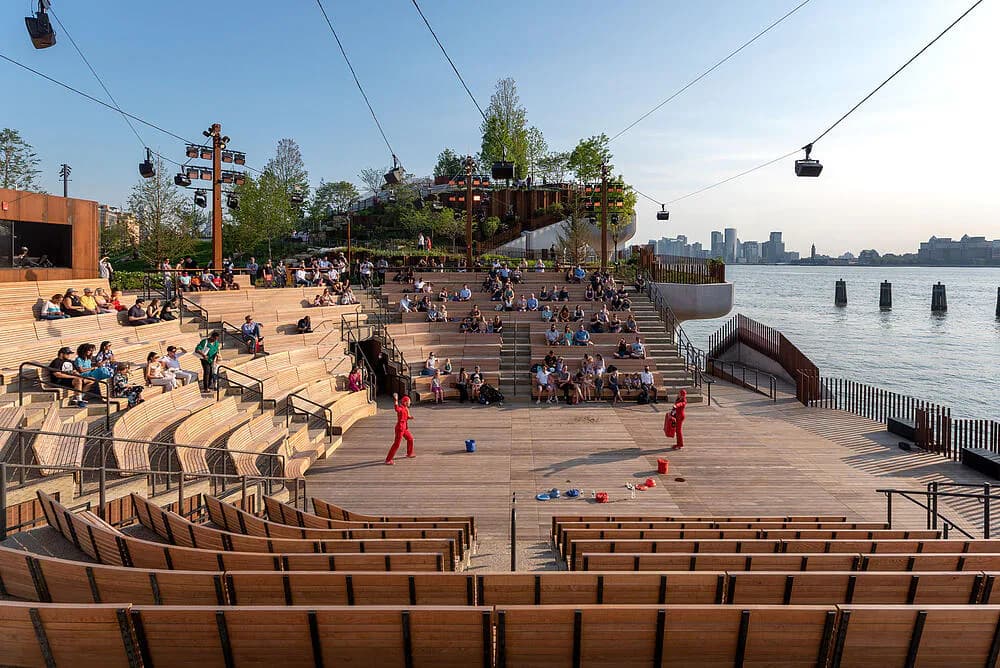 Waterfront plaza with tiered wooden amphitheater seating, sandy beach areas, and water views, featuring public gathering spaces and landscape design by SITU.