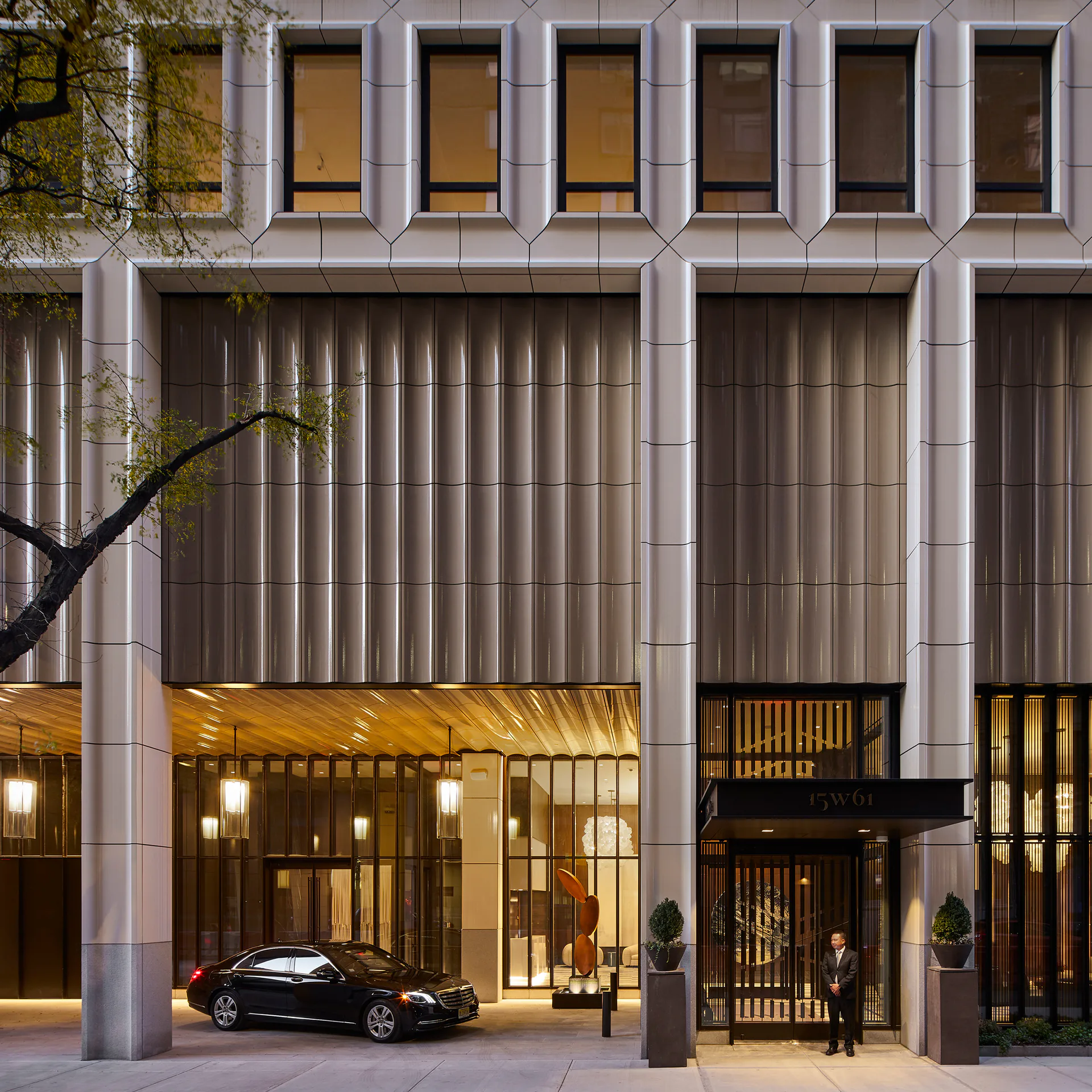 Modern luxury building facade with polished stone and vertical metal paneling, illuminated storefront with gold lighting and black car parked at entrance, by Skidmore, Owings & Merrill.