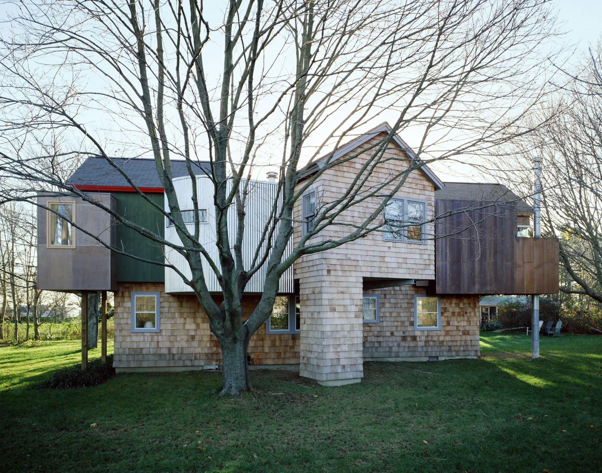 Modern residential structure with stone and wood cladding elevated on pillars, featuring angular roof forms and multiple protruding volumes, surrounded by bare trees and green lawn, by SKOLNICK Architecture.