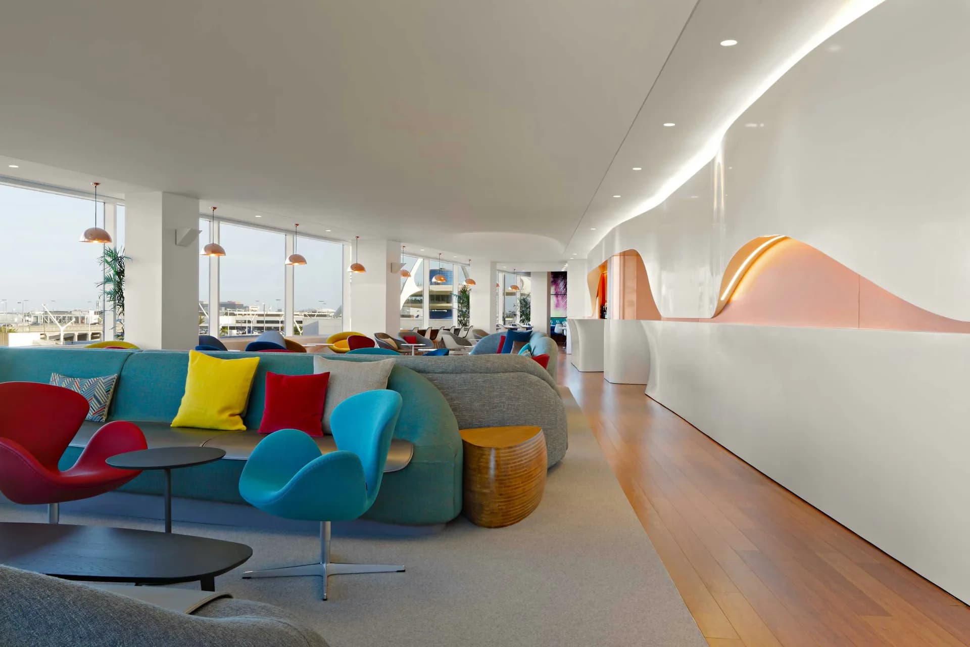 Bright modern lounge space with colorful modular seating, suspended pendant lights, and orange sculptural wall elements overlooking coastal views by SLADE ARCHITECTURE.