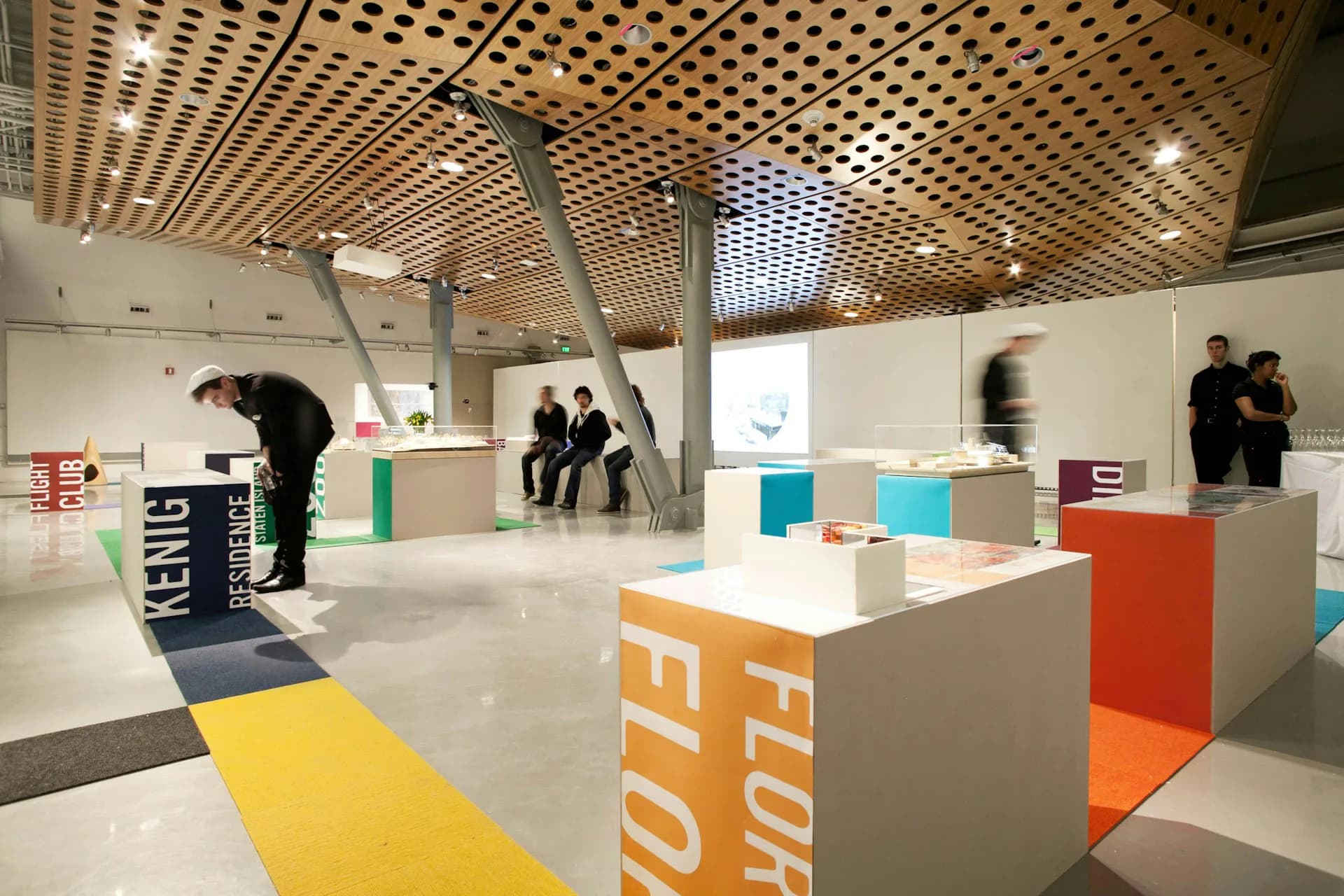 A modern exhibition space with wooden lattice ceiling, colorful floor pathways in yellow and orange, and white display cubes with typography by SLADE ARCHITECTURE.