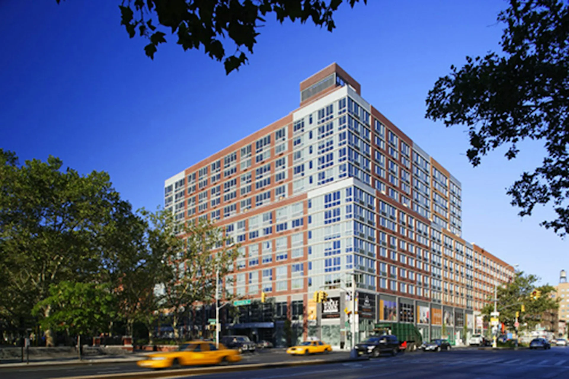 Brick and glass residential building with orange and brown facades, ground-floor retail, trees, and yellow taxi on street by SLCE Architects, LLP.