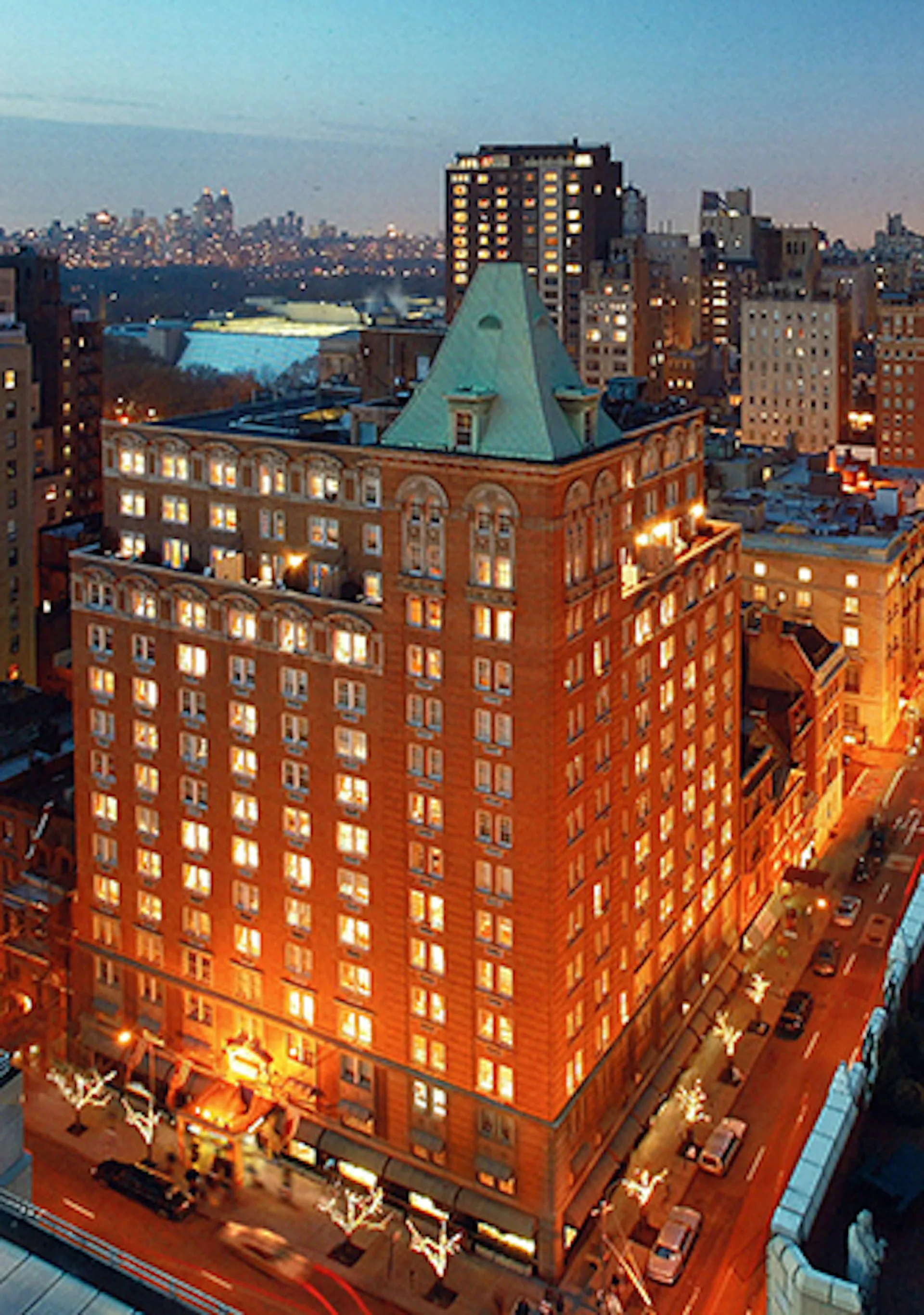 Illuminated brick and copper-roofed historic building at dusk with Manhattan skyline in background, designed by SLCE Architects, LLP.