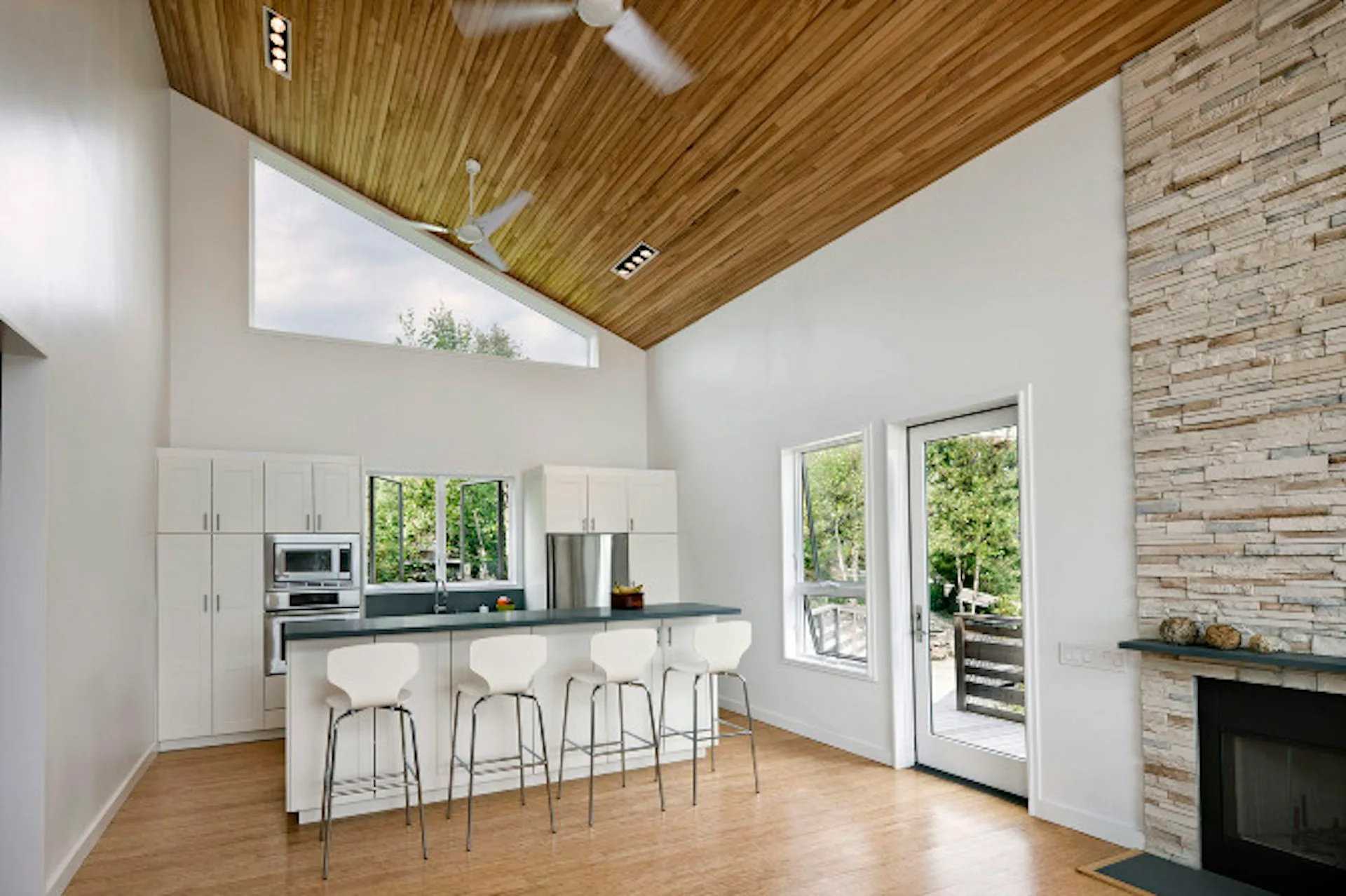 Modern kitchen and living space with wood ceiling, white cabinetry, island seating, brick fireplace, and glass doors by Smith and Architects.