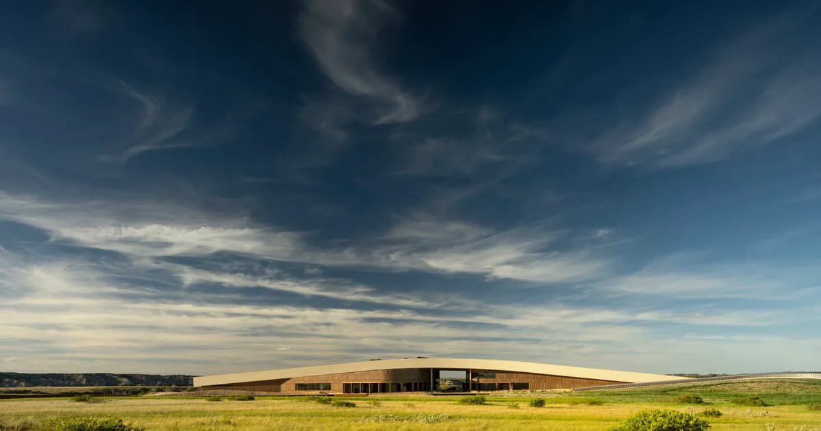 A modern low-rise agricultural building with dark metal cladding sits across an expansive flat landscape under a dramatic cloudy sky, designed by Snøhetta New York.