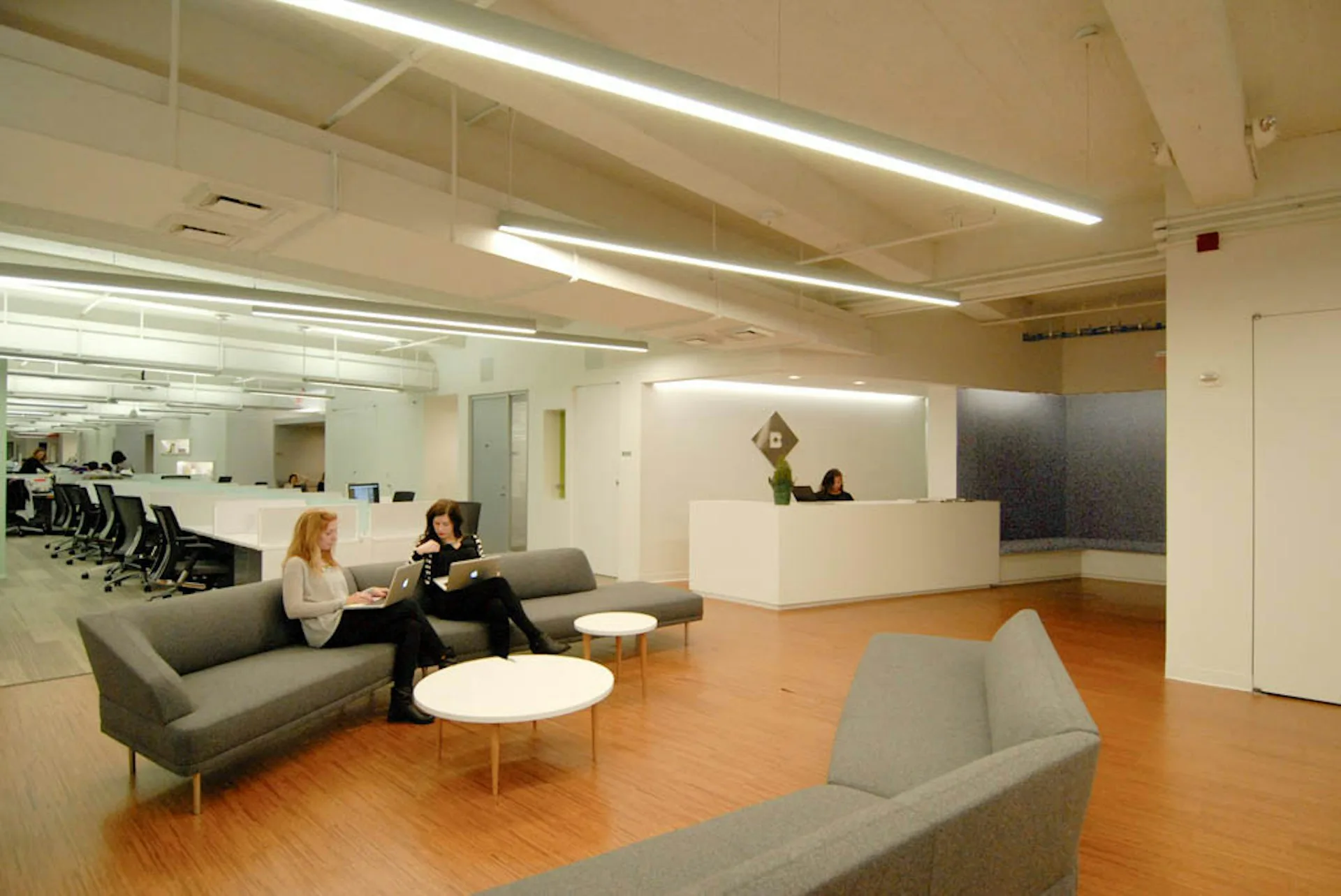 Modern office lobby with gray upholstered seating, white reception desk, orange wood flooring, and glass partitions separating open workspaces by Soluri Architecture.