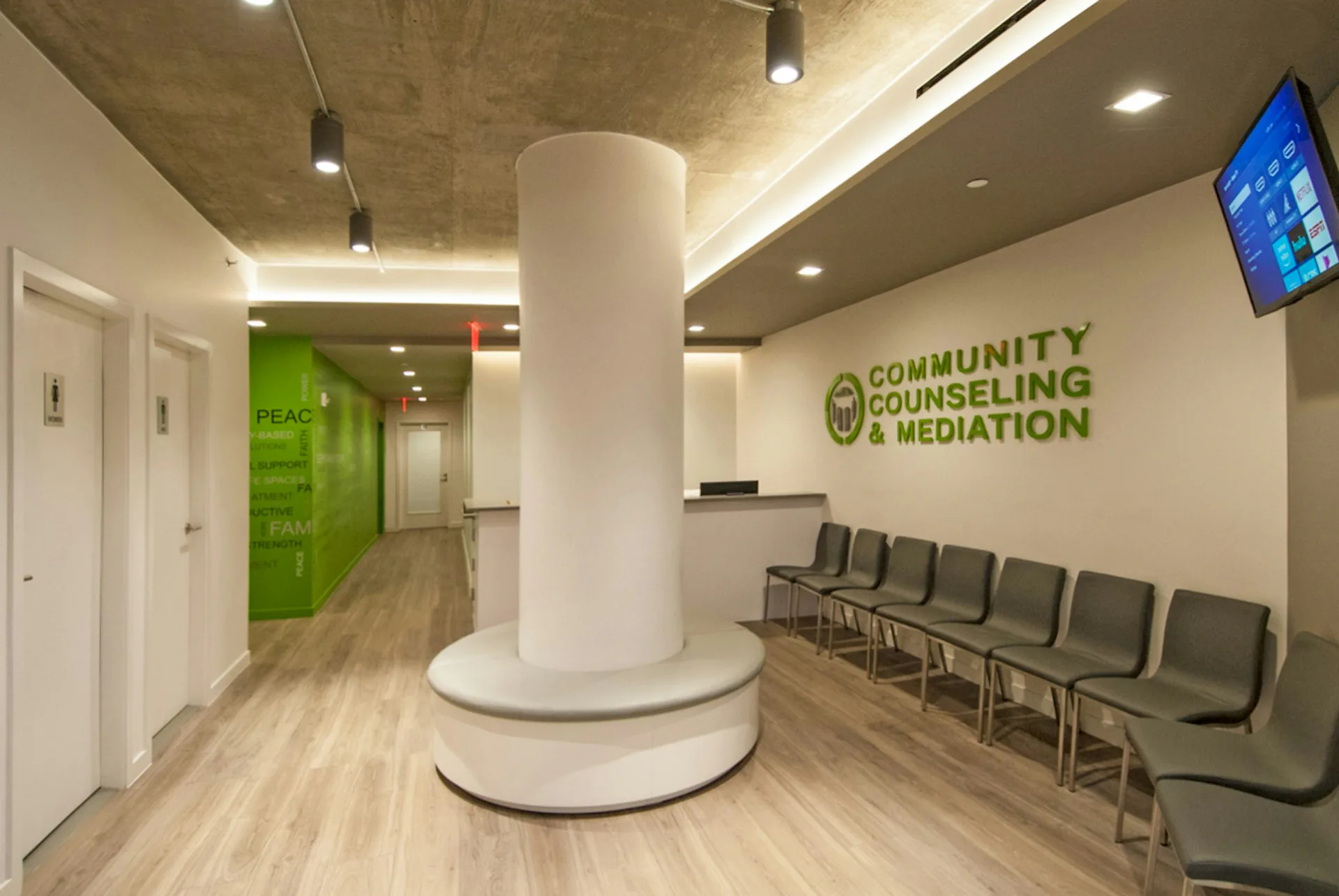 Community Counseling & Mediation office reception area with cylindrical white column, green accent wall, wood flooring, and gray seating by Soluri Architecture.