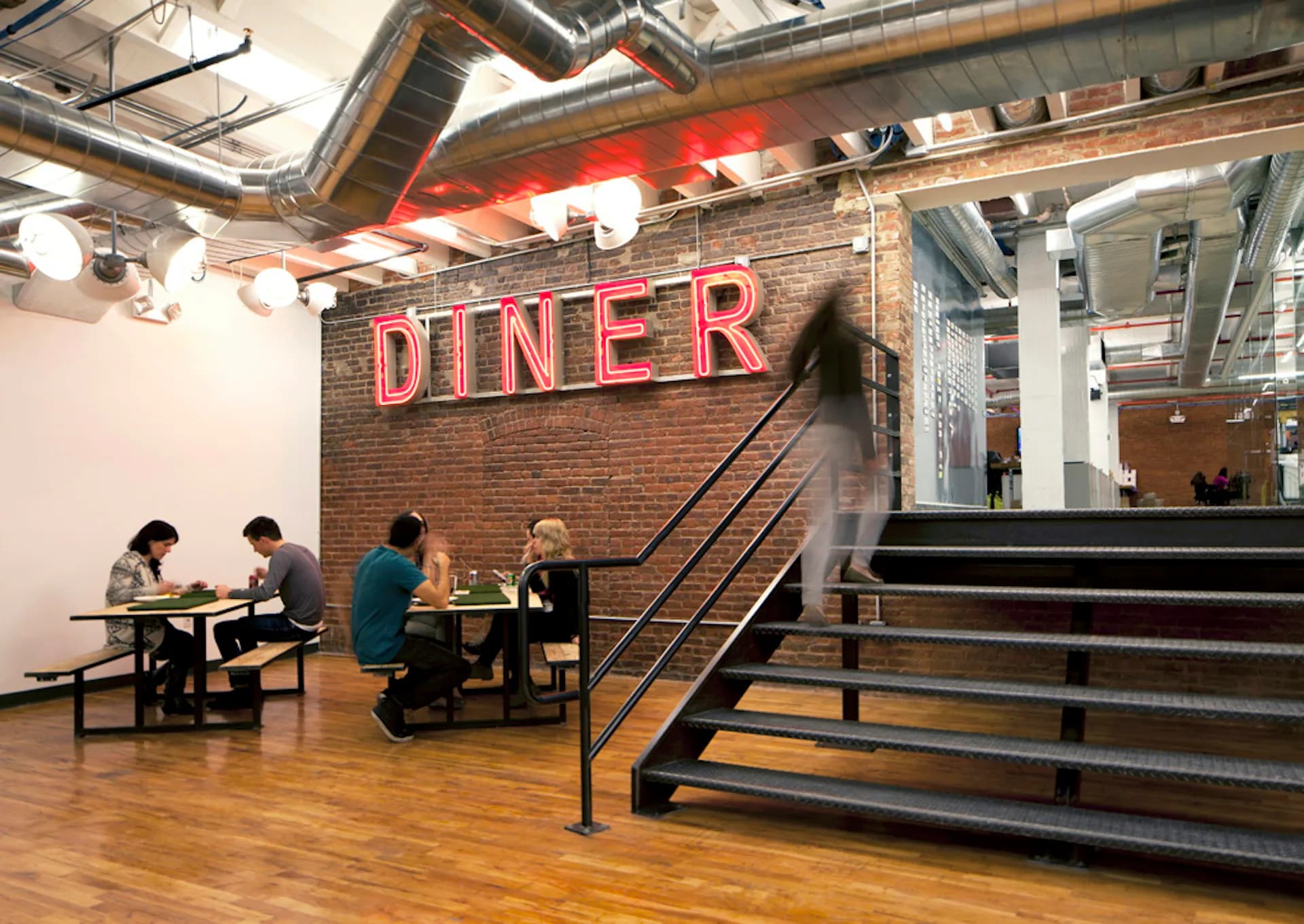 Industrial loft dining space with exposed brick, neon 'DINER' signage, wooden floors, metal stairs, and industrial ceiling elements by Spectorgroup.