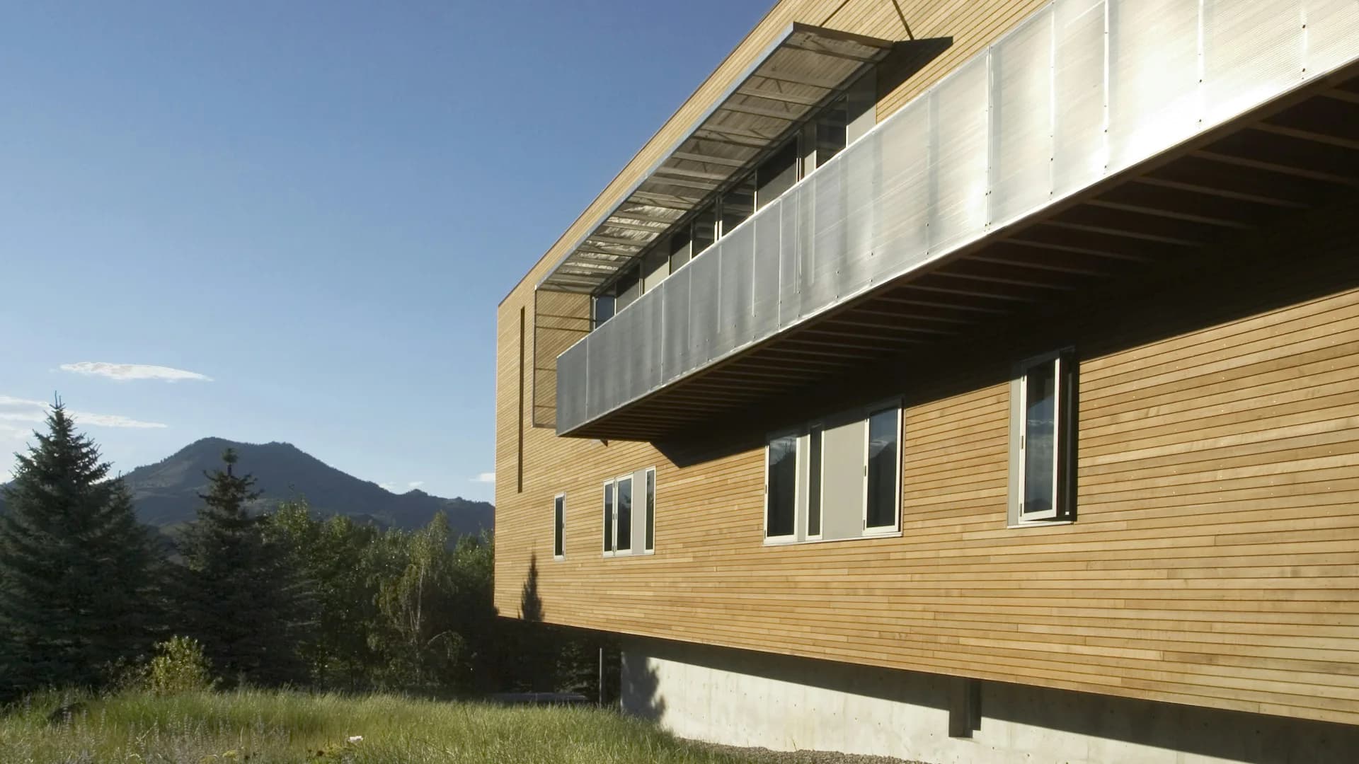 Modern residential building with natural wood siding and metal upper level, positioned against forested mountain backdrop by SPG Architects.