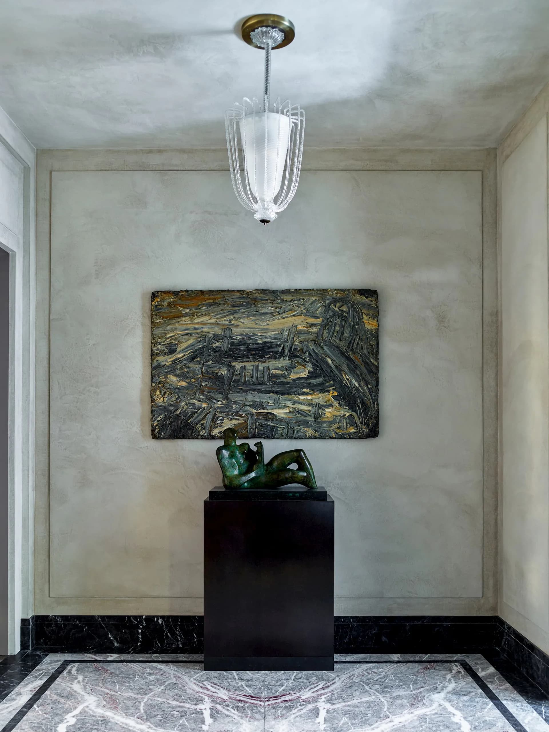 A minimalist gallery entry featuring polished marble flooring, concrete walls, a black pedestal with sculptural object, abstract artwork, and modern pendant light by S.R. Gambrel.