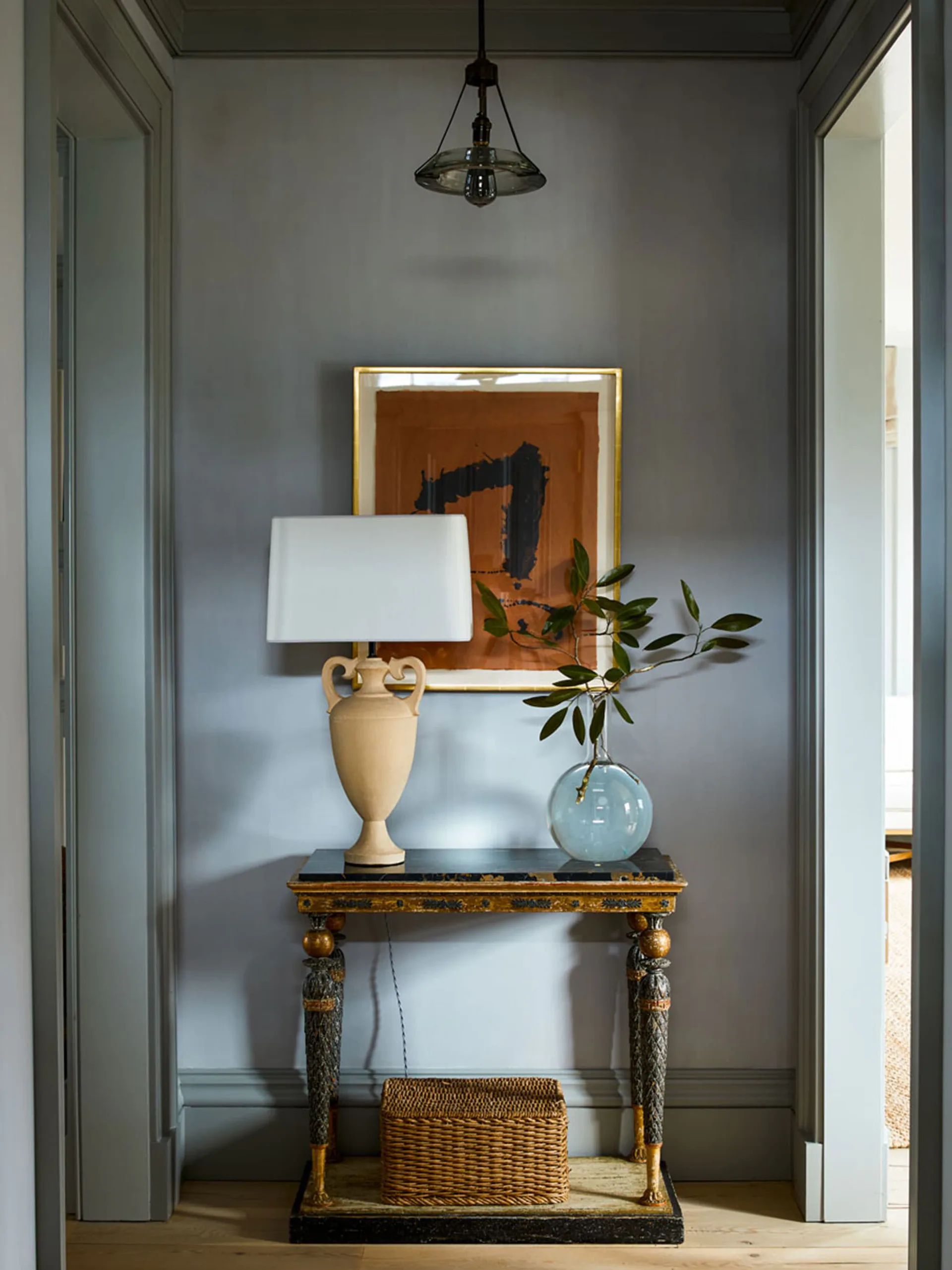 A hallway alcove with gray walls features a gilt console table displaying a ceramic lamp, glass vase with greenery, and woven basket, beneath framed artwork and a pendant light by S.R. Gambrel.