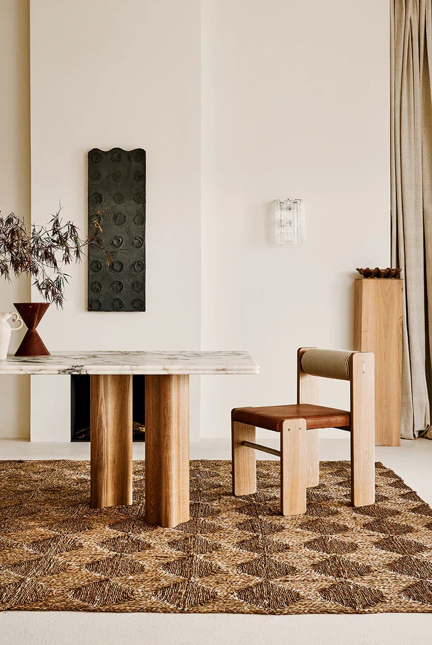 Minimalist dining space with wooden table and chair on natural fiber rug, white walls, and dark sculptural wall art by SSS Atelier.